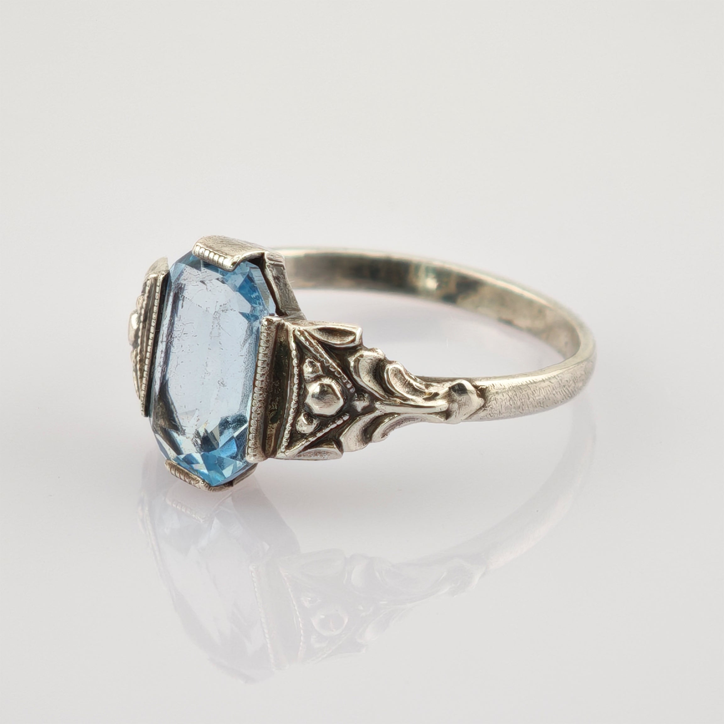 Antiker Silberring mit dekorativem Band und facettiertem blauem Edelstein auf reflektierender Oberfläche.