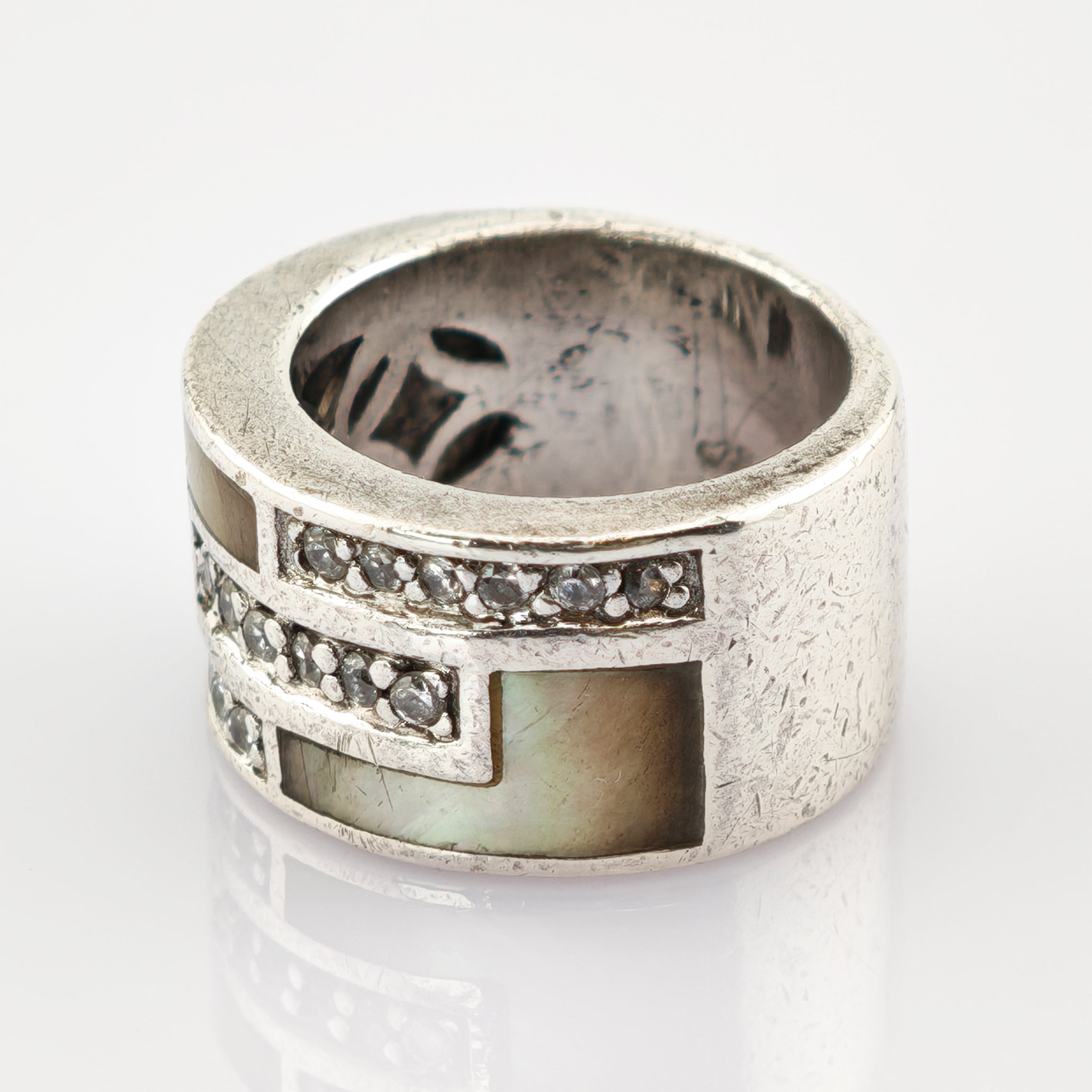 Breiter Silberring mit geometrischen Einlagen aus Perlmutt und kleinen klaren Steinen auf reflektierender Oberfläche