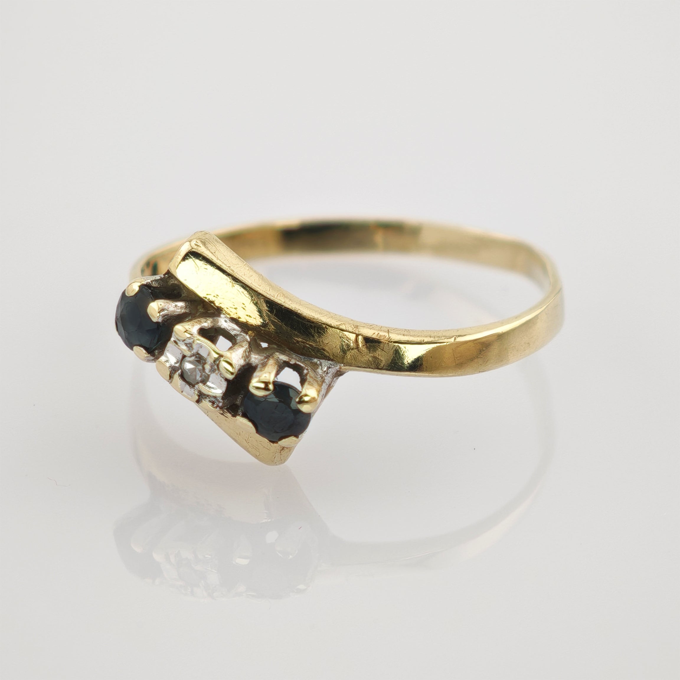 Goldring mit asymmetrischem Band und drei Steinen, zwei dunkelblau und einem klaren, auf weißem Eleganter Goldring mit gebogenem Steinbesatz aus zwei dunkelblauen und einem weißen Edelstein.