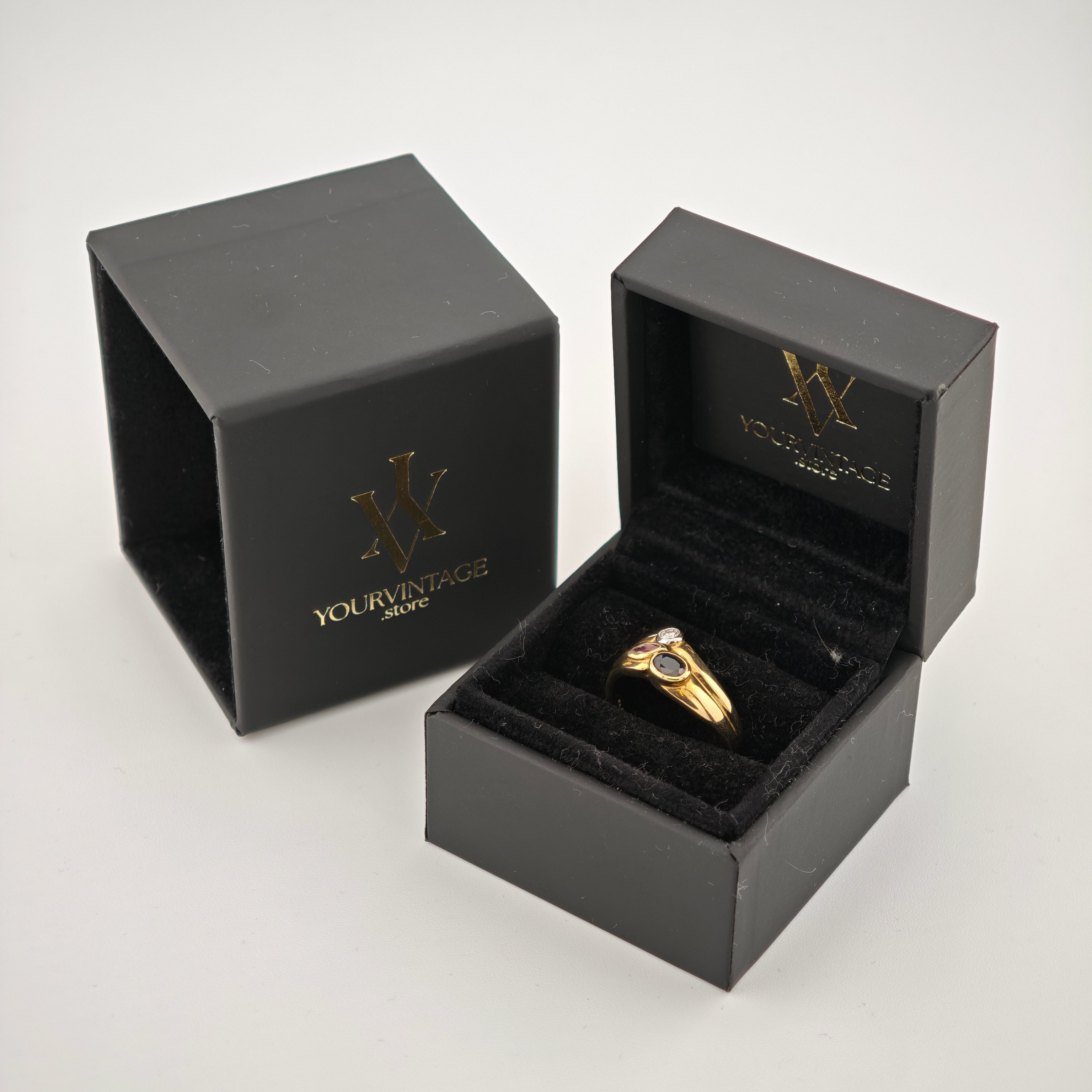 Geöffnete schwarze Schmuckboxen mit Goldlogo und goldenem Ring auf schwarzem Samt.