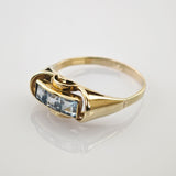 Goldring mit drei quadratischen hellblauen Edelsteinen in Goldener Ring mit rechteckigem Oberteil und drei blauen quadratischen Edelsteinen auf reflektierendem Untergrund.