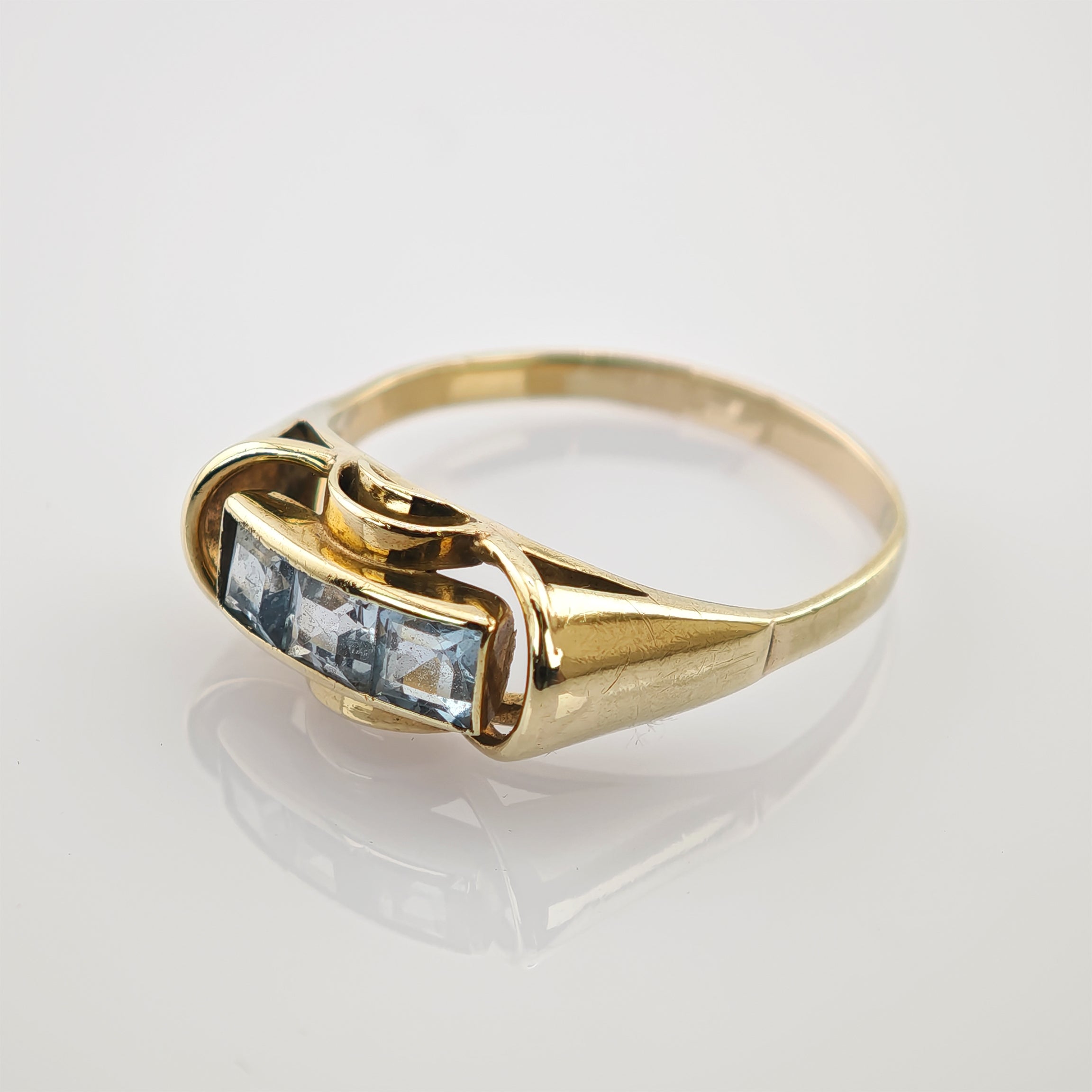 Goldring mit drei quadratischen hellblauen Edelsteinen in Goldener Ring mit rechteckigem Oberteil und drei blauen quadratischen Edelsteinen auf reflektierendem Untergrund.