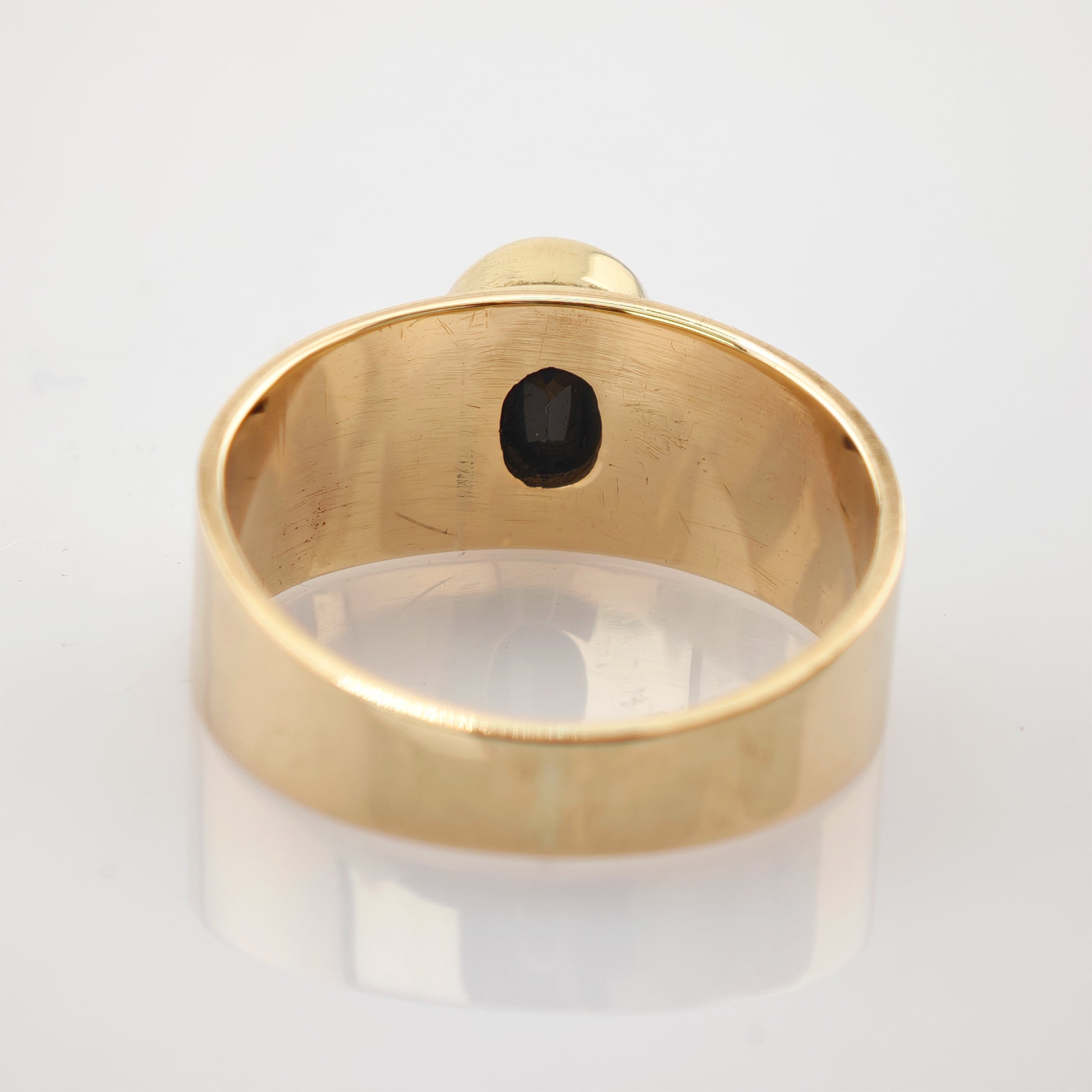 Goldener Ring mit breitem Band und dunklem ovalem Inset auf reflektierender Oberfläche.