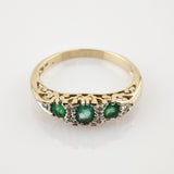 Goldener Ring mit filigraner Gravur und drei horizontal angeordneten Smaragden, umgeben von kleinen weißen Steinen.