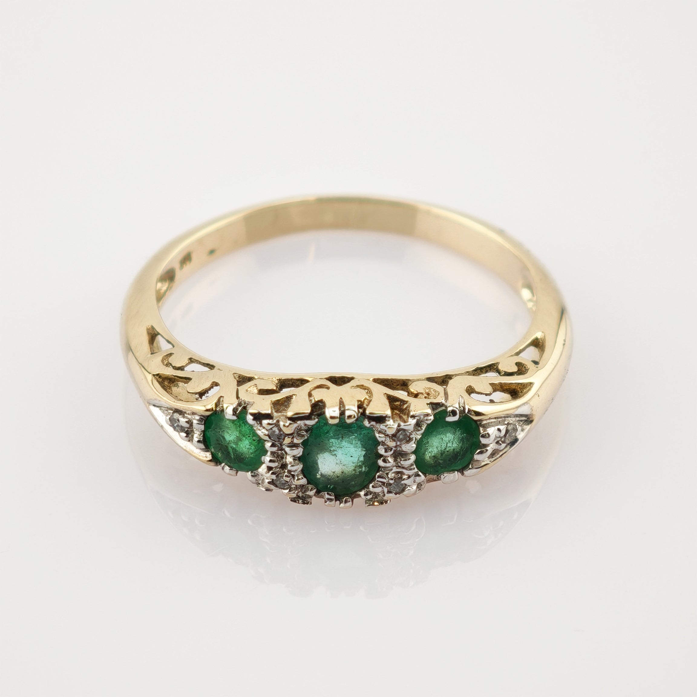 Goldener Ring mit filigraner Gravur und drei horizontal angeordneten Smaragden, umgeben von kleinen weißen Steinen.