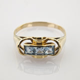 Goldener Ring mit drei quadratischen hellblauen Edelsteinen und geschwungenen Details auf weißem Goldener Ring mit drei quadratisch geschliffenen hellblauen Edelsteinen in Reihe