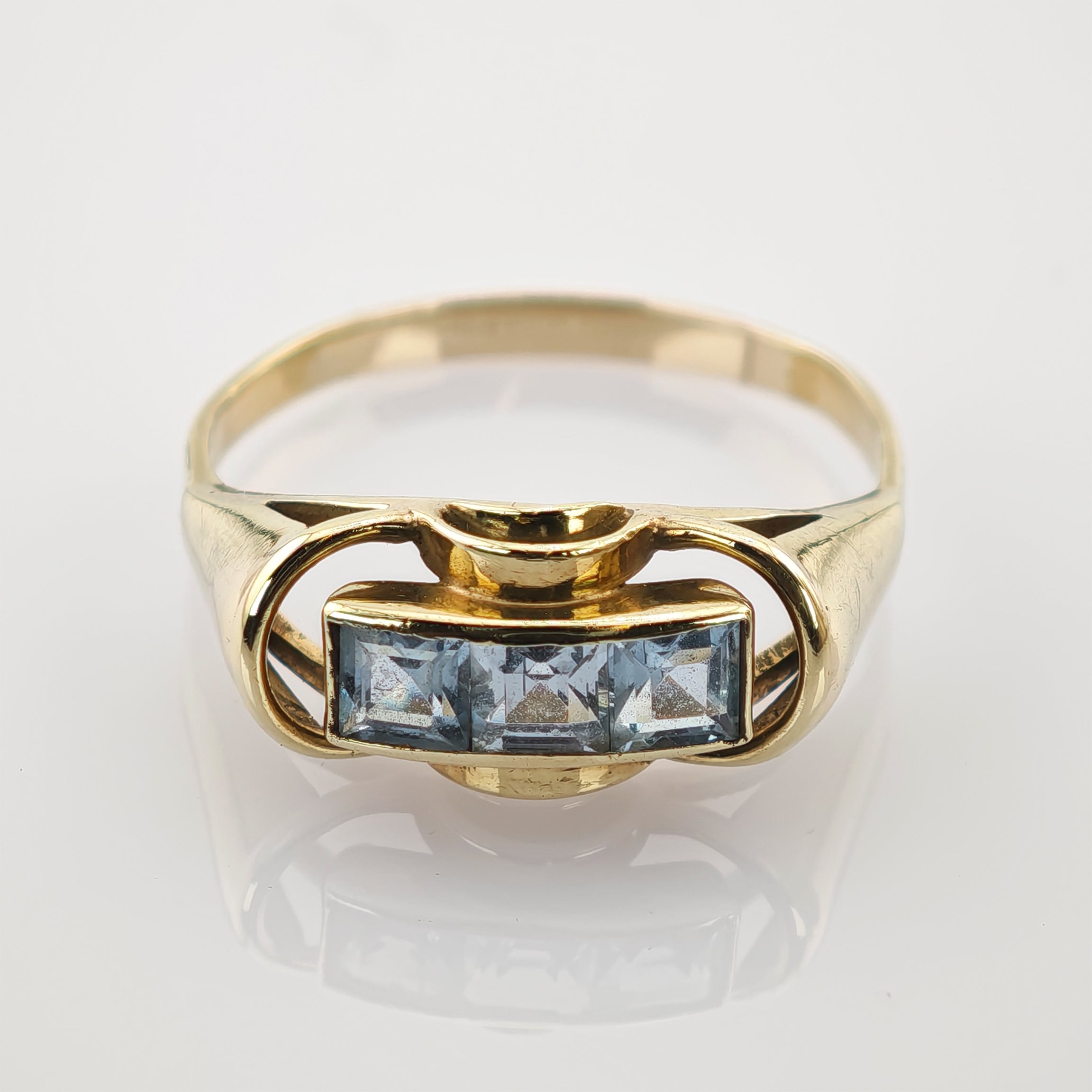 Goldener Ring mit drei quadratischen hellblauen Edelsteinen und geschwungenen Details auf weißem Goldener Ring mit drei quadratisch geschliffenen hellblauen Edelsteinen in Reihe