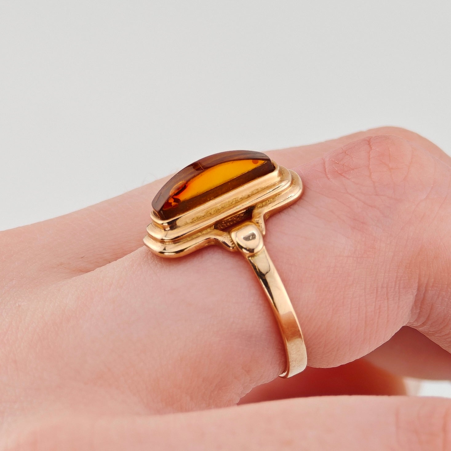 Goldener Ring mit ovalem bernsteinfarbenem Stein und glatter Bandstruktur, an einer Hand vor hellem Hintergrund.