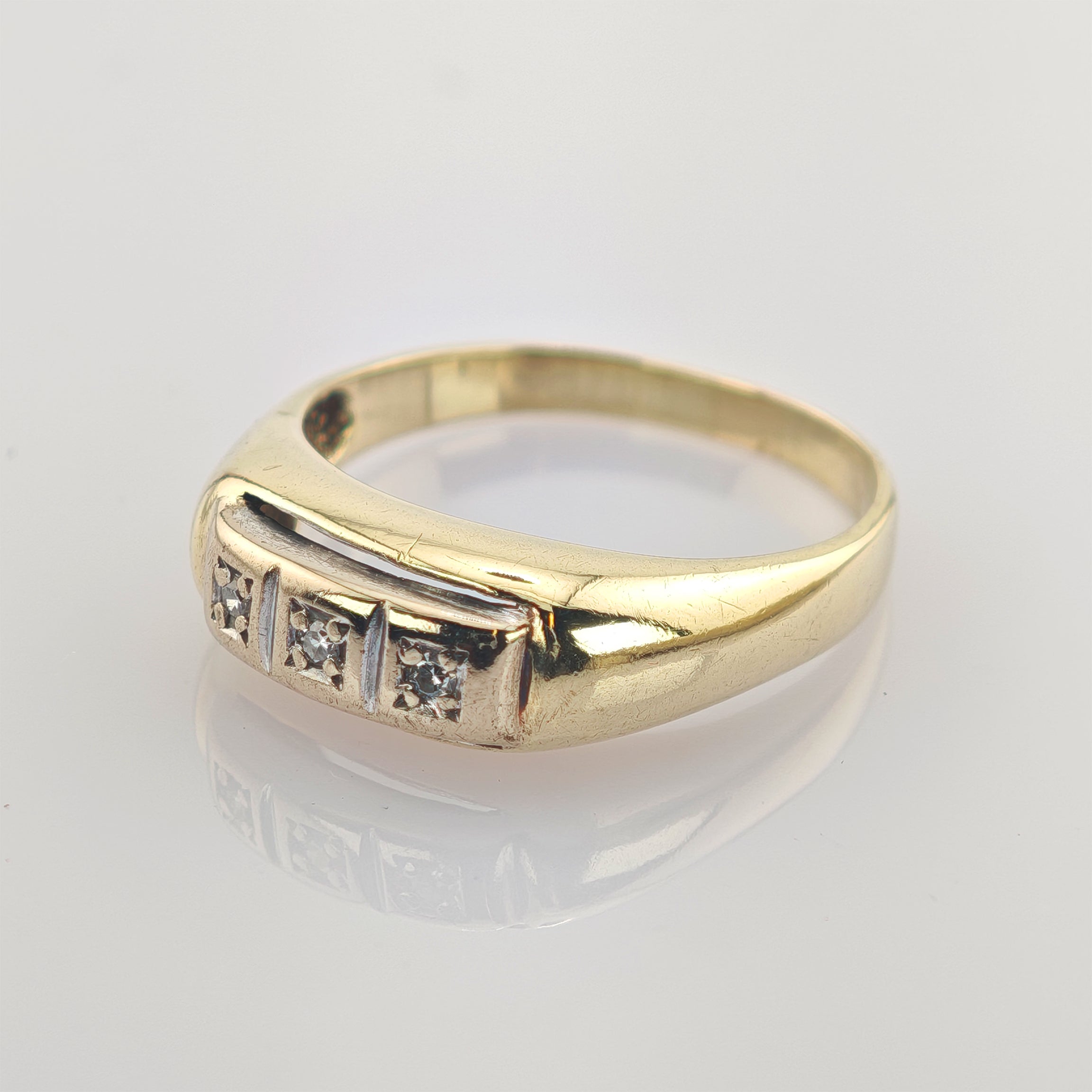 Goldener Ring mit poliertem Band und drei kleinen Diamanten in quadratischen Fassungen auf weißer reflektierender Oberfläche.