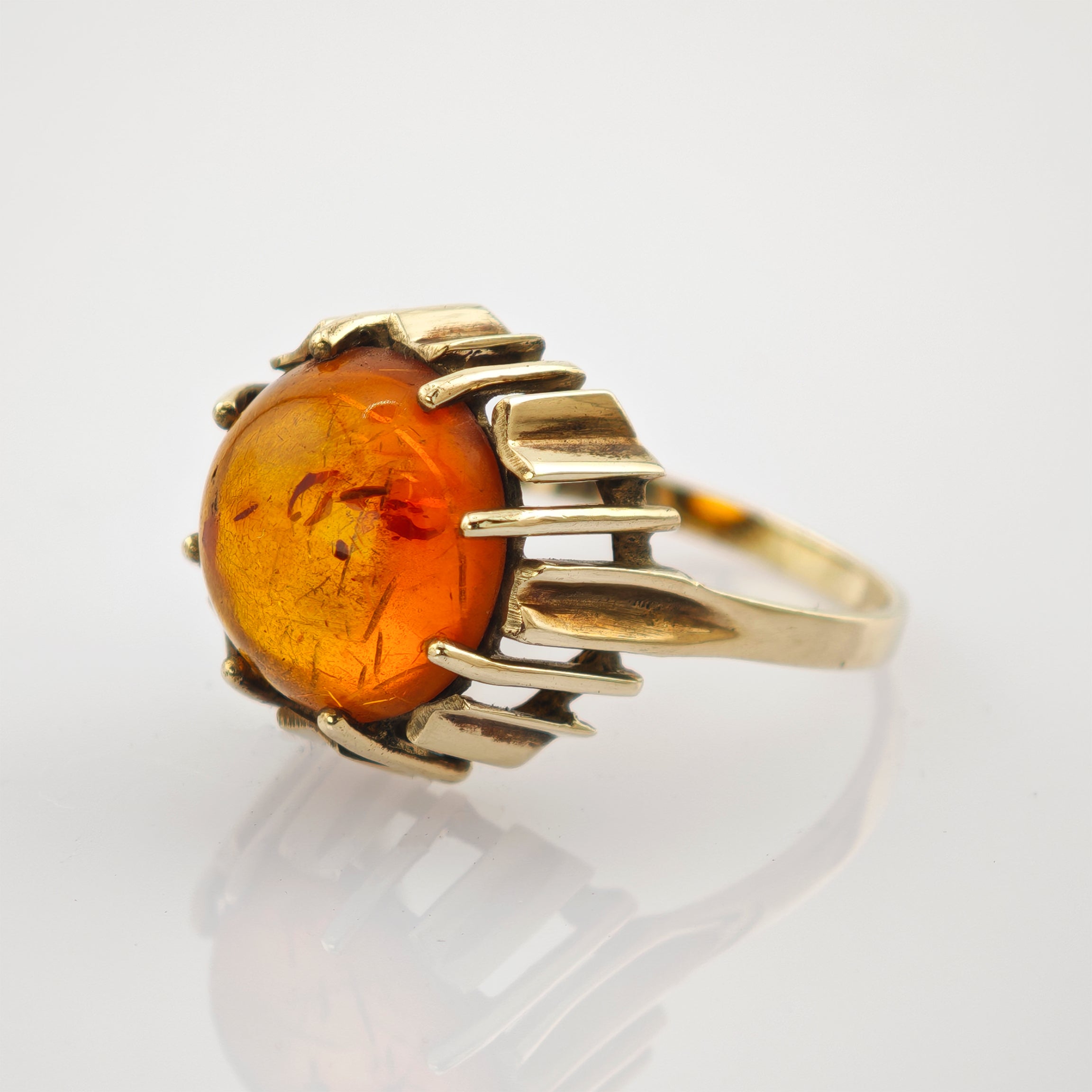 Goldener Ring mit rundem Bernstein in geometrischer Fassung auf weißem, reflektierendem Untergrund.