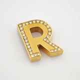 Goldener Schmuckbuchstabe R mit runden Edelsteinen auf weißem Hintergrund.