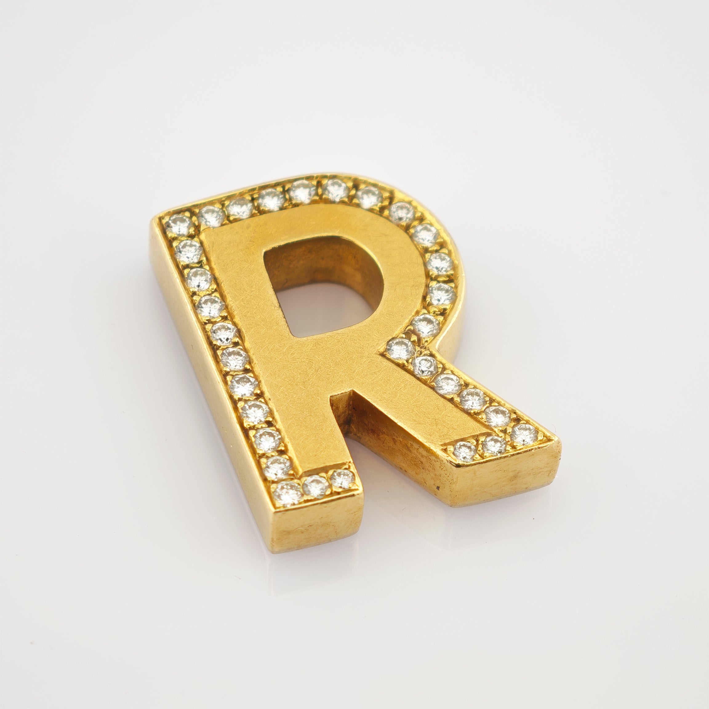 Goldener Schmuckbuchstabe R mit runden Edelsteinen auf weißem Hintergrund.
