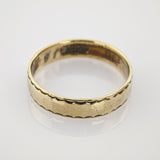 Goldener Hochzeitsring mit gezacktem Muster auf spiegelnder weißer Goldring mit einfacher geschwungener Musterkante und leichten Gebrauchsspuren auf weißer Oberfläche