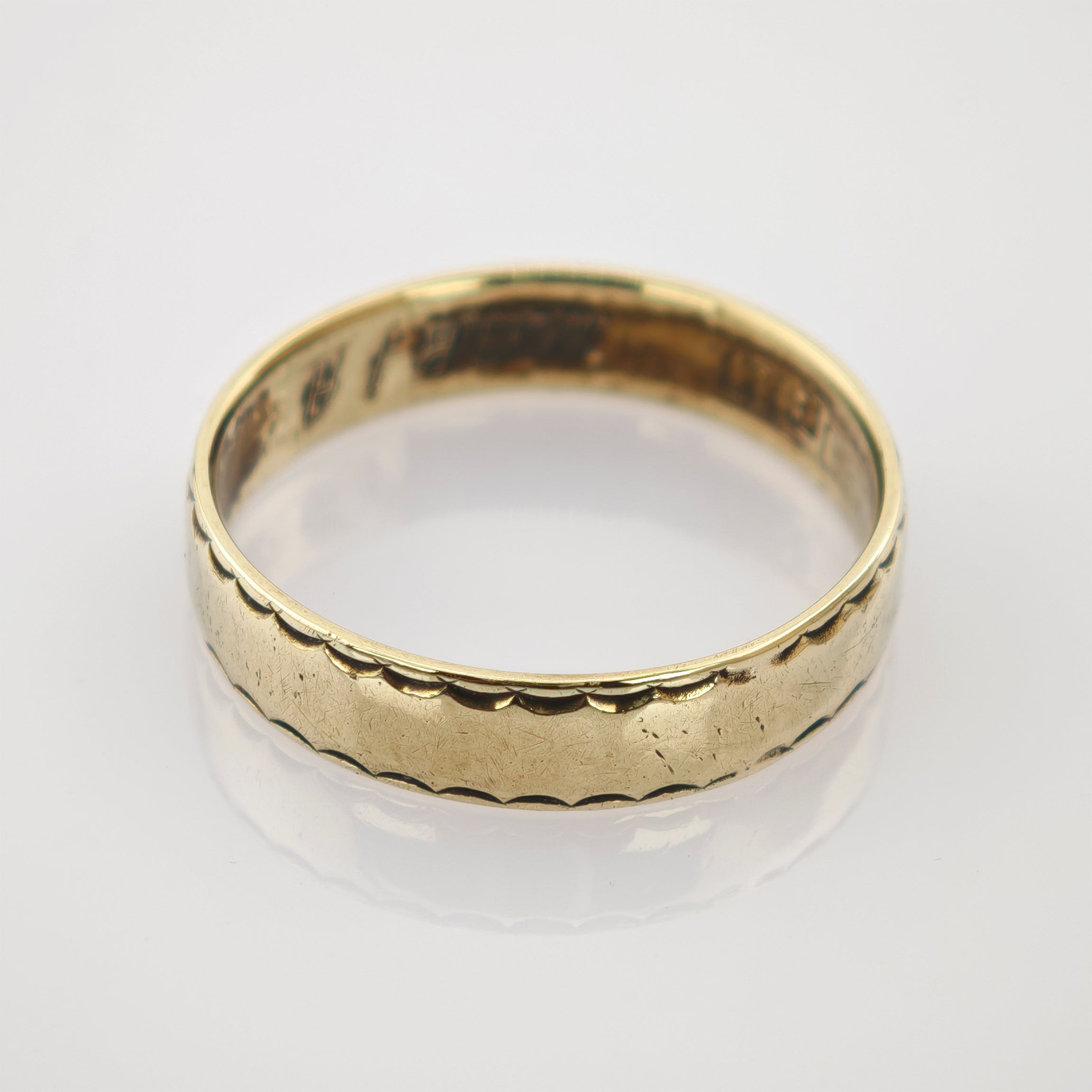 Goldener Hochzeitsring mit gezacktem Muster auf spiegelnder weißer Goldring mit einfacher geschwungener Musterkante und leichten Gebrauchsspuren auf weißer Oberfläche