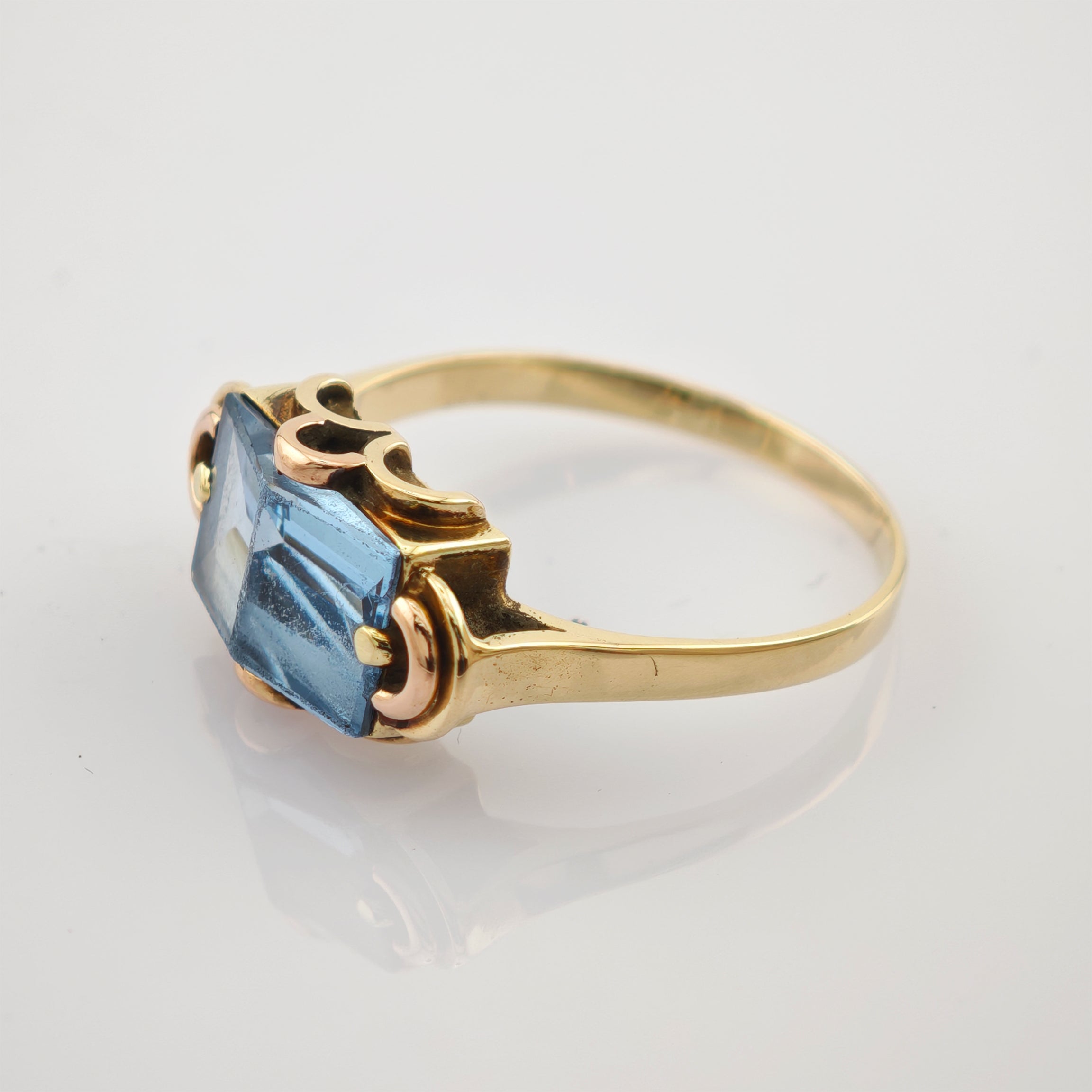 Goldring mit quadratischem blauem Edelstein und dekorativen Goldelementen auf weißem Hintergrund.