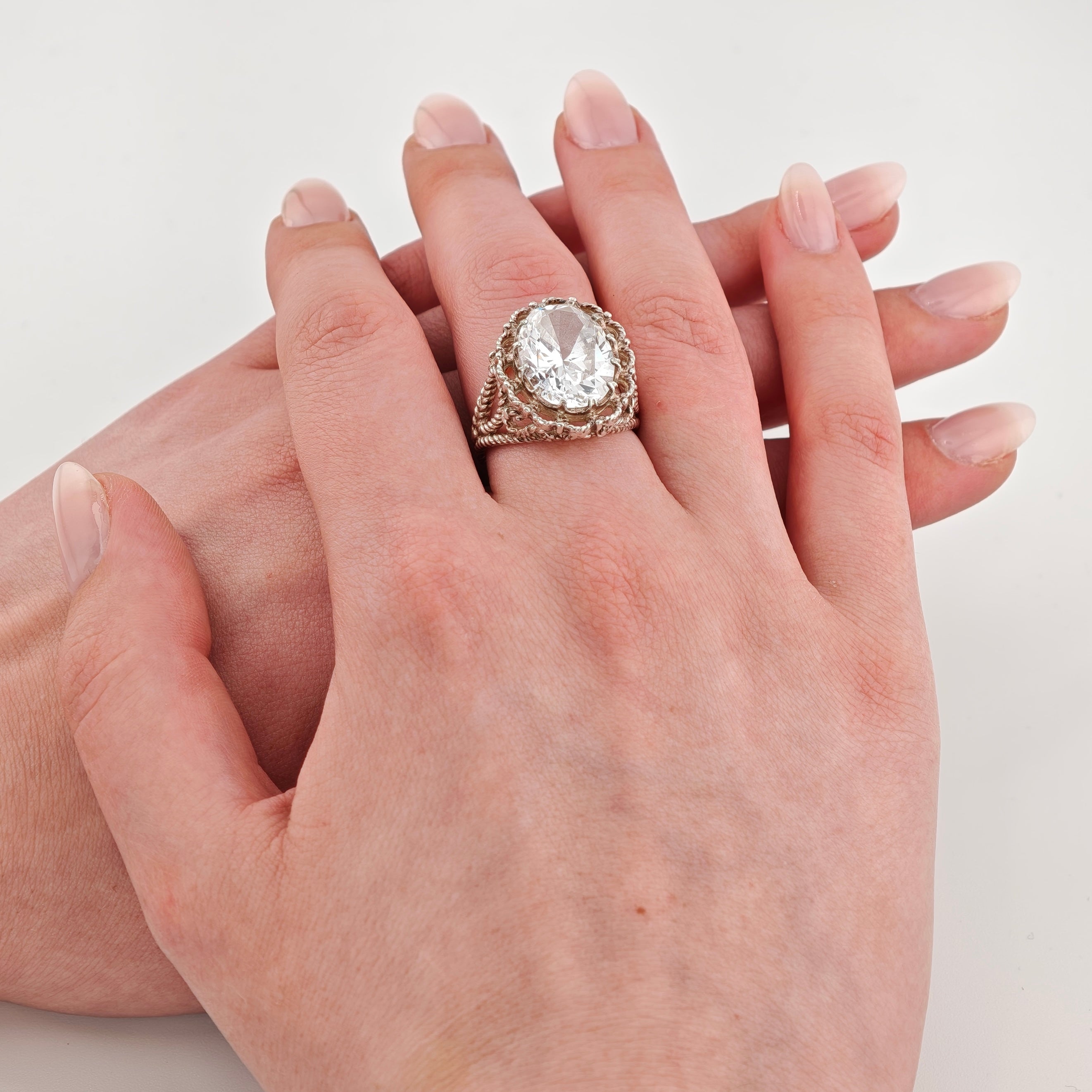 Zwei helle Hände, obere Hand trägt einen silbernen Ring mit gedrehtem Band und großem ovalem klarem Edelstein, gepflegte Nägel mit hellem rosa Nagellack.