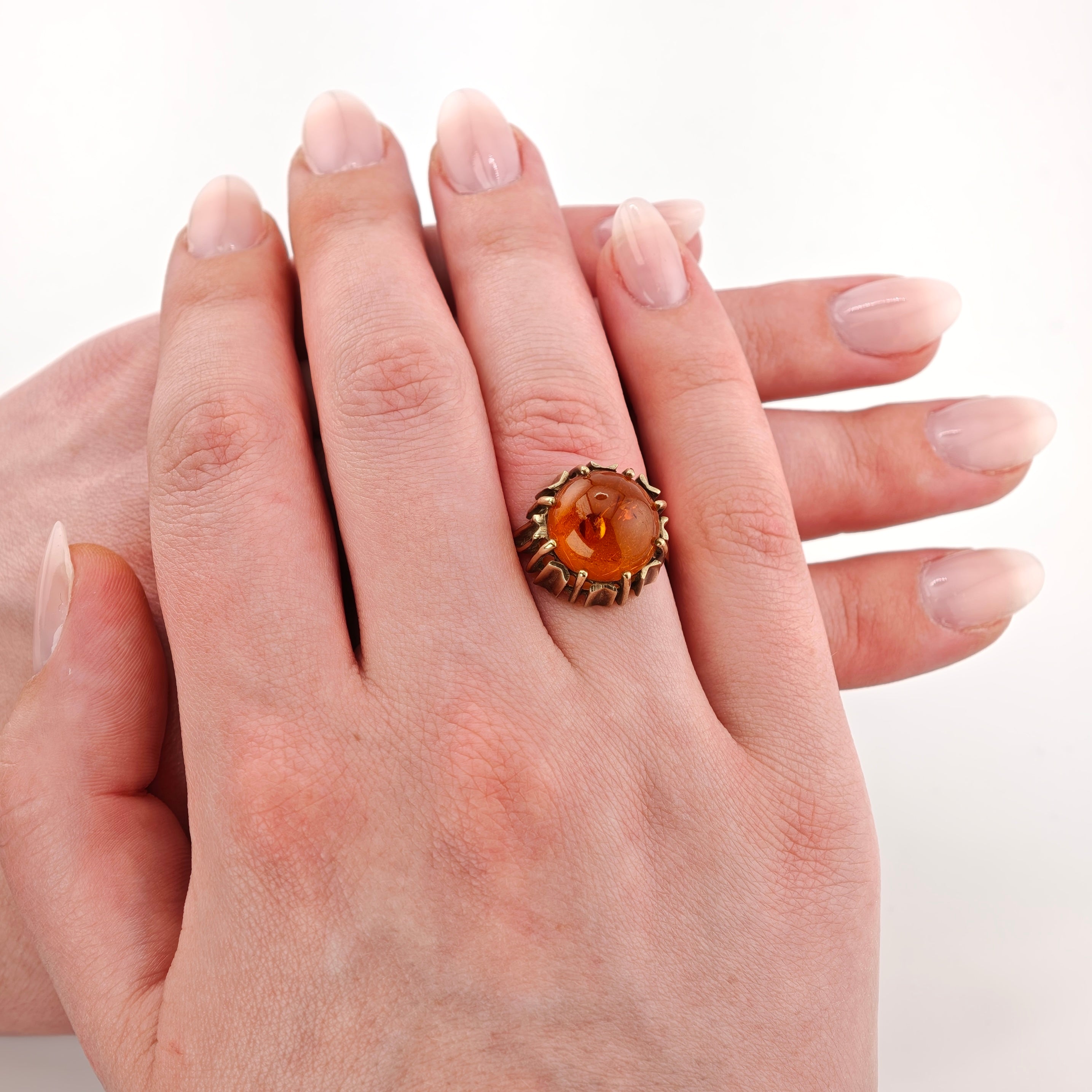 Hand mit Goldring und rundem Bernsteinstein am Mittelfinger vor weißem Hintergrund, elegant manikürt.