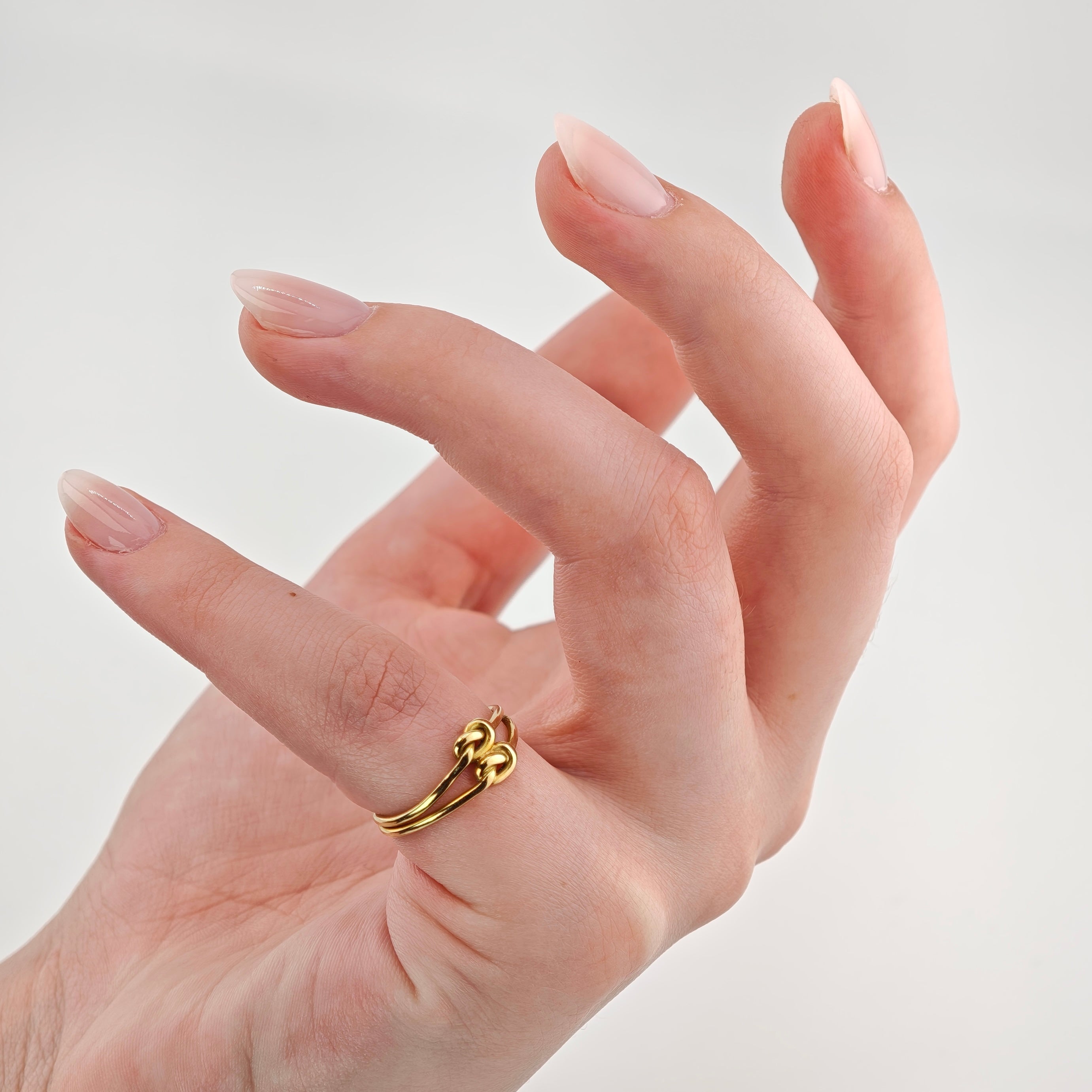 Hand mit hellrosa lackierten Nägeln trägt goldenen Ring mit Doppelknoten-Design am Mittelfinger.