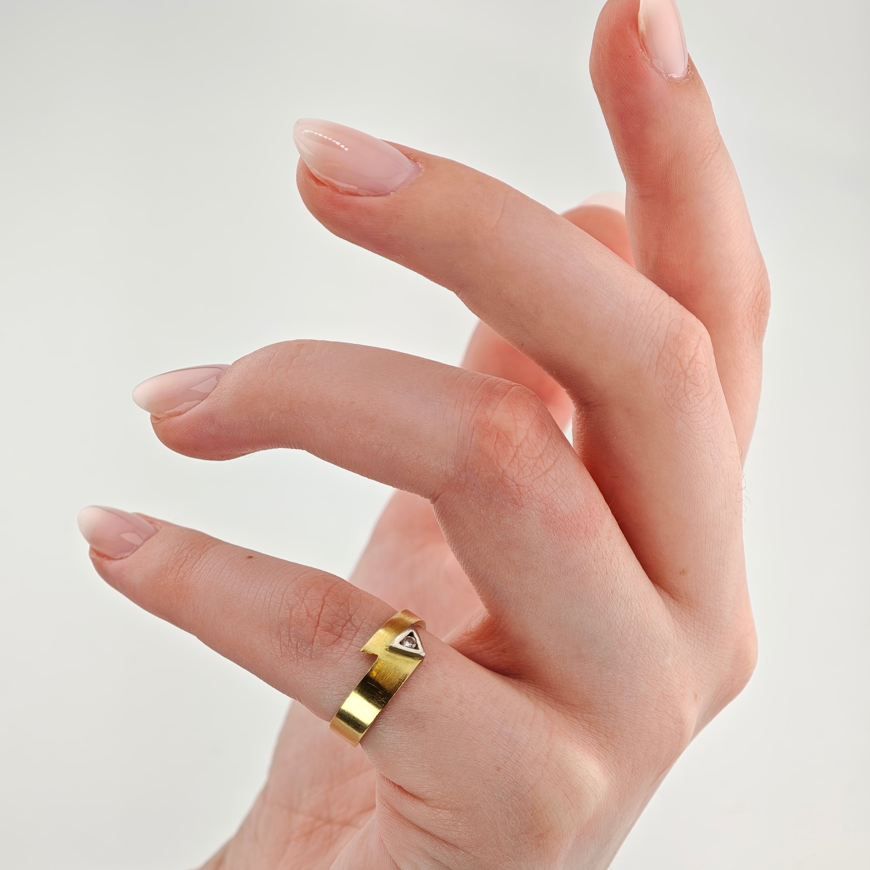 Nahaufnahme einer Hand mit hellrosa lackierten Nägeln und einem goldenen Ring mit dreieckigem Diamanten.