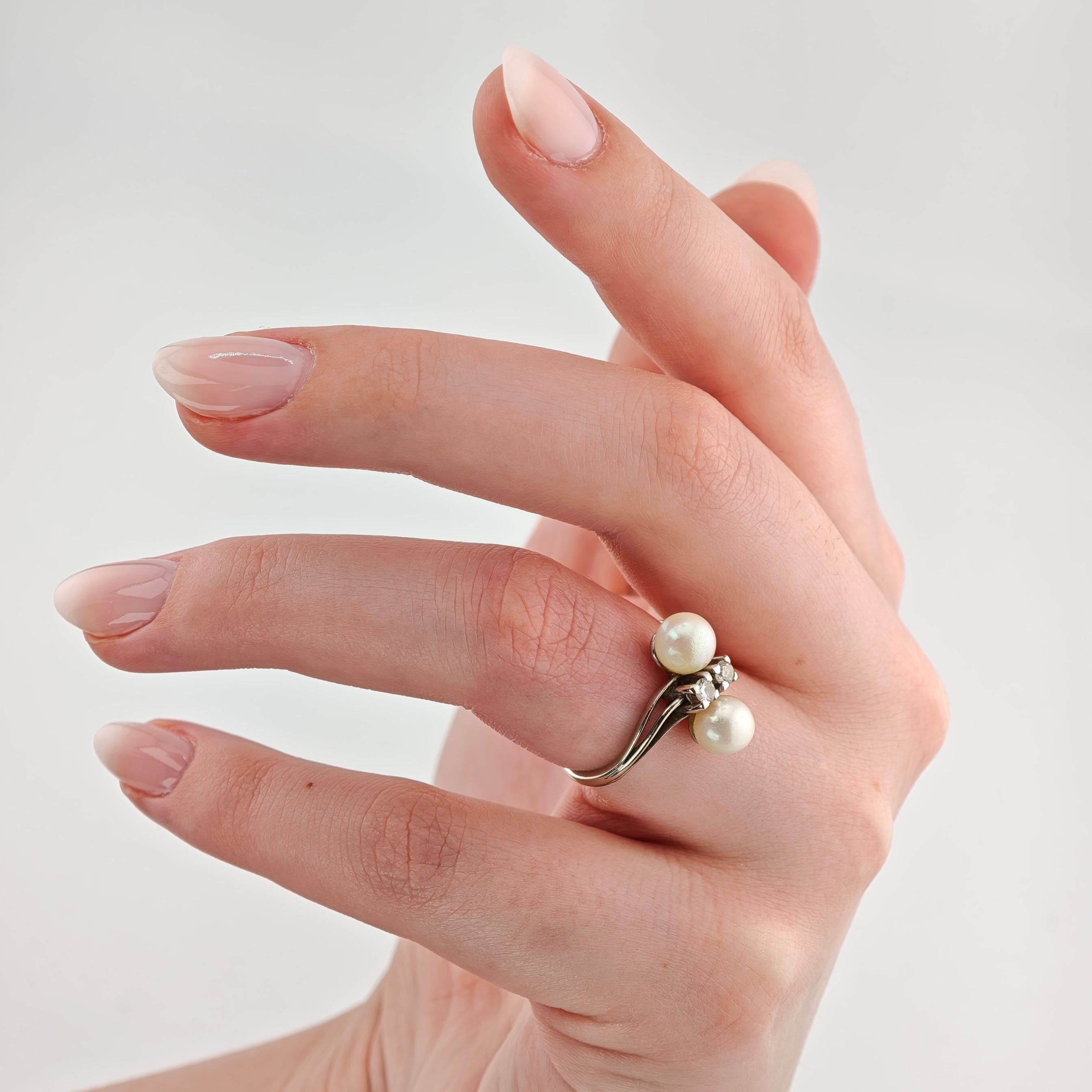 Hand mit elegantem Silberring mit zwei weißen Perlen und kleinen klaren Steinen auf rosa Nude-Lack.