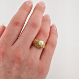Linke Hand mit einem goldenen Ring mit Goldschuppen und einer weißen Perle vor hellem Hintergrund.