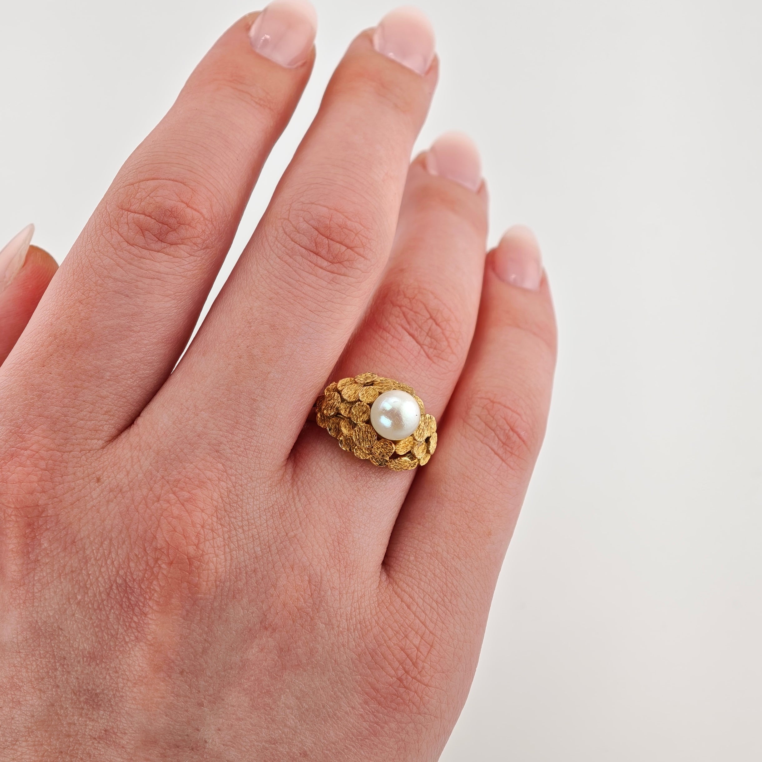 Linke Hand mit einem goldenen Ring mit Goldschuppen und einer weißen Perle vor hellem Hintergrund.