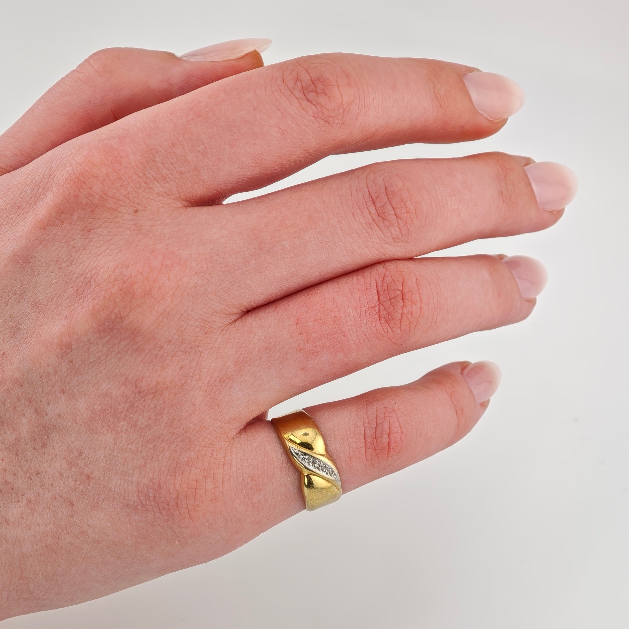 Linke Hand einer hellhäutigen Person trägt einen goldenen Ring mit wellenförmigem Design und kleinen Edelsteinen.
