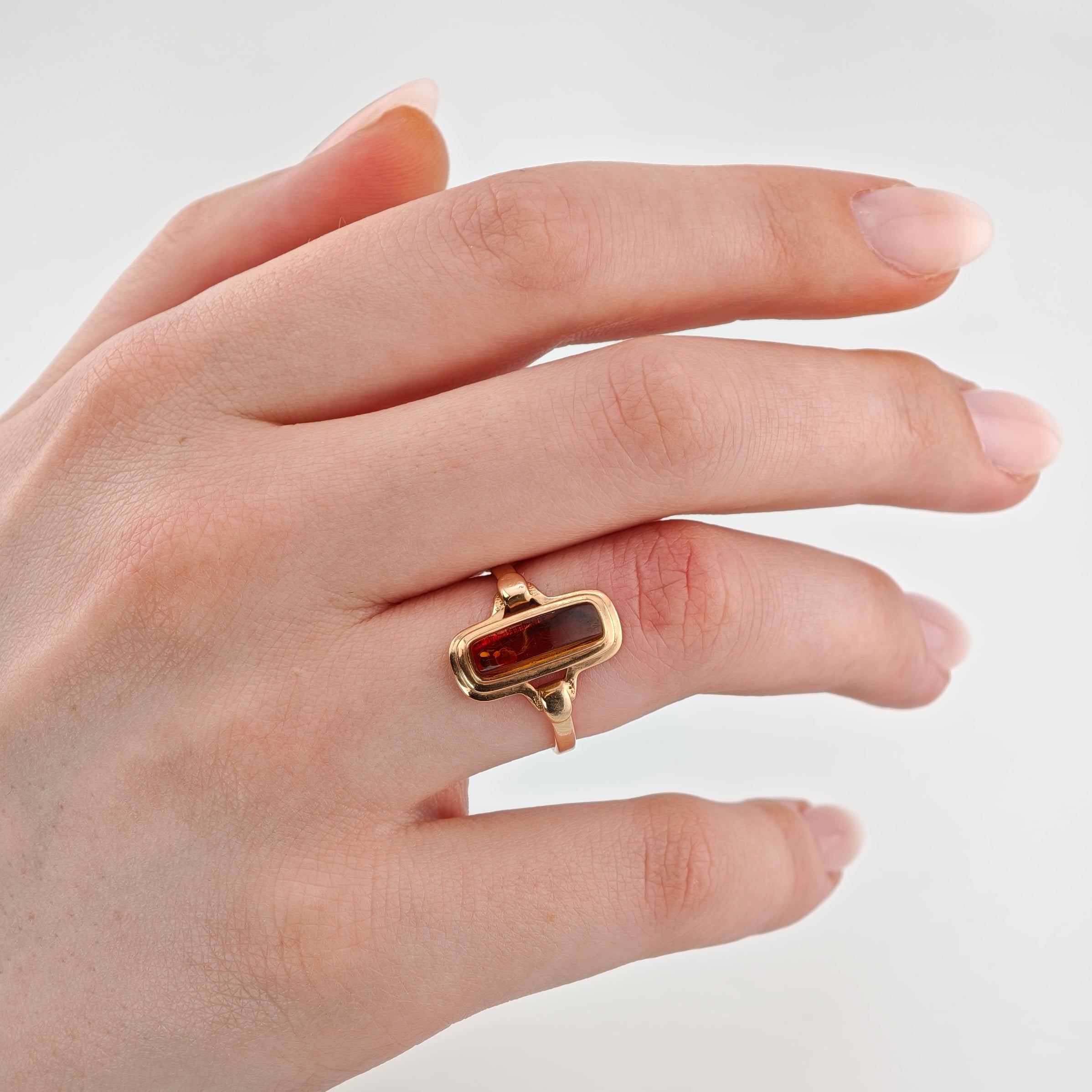 Linke Hand mit Goldring und rechteckigem Bernstein auf dem Ringfinger, hellrosa Maniküre, weißer Hintergrund.