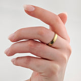 Linke Hand mit rosa lackierten Nägeln und goldfarbenem Ring mit gewellten Kanten vor neutralem Hintergrund.