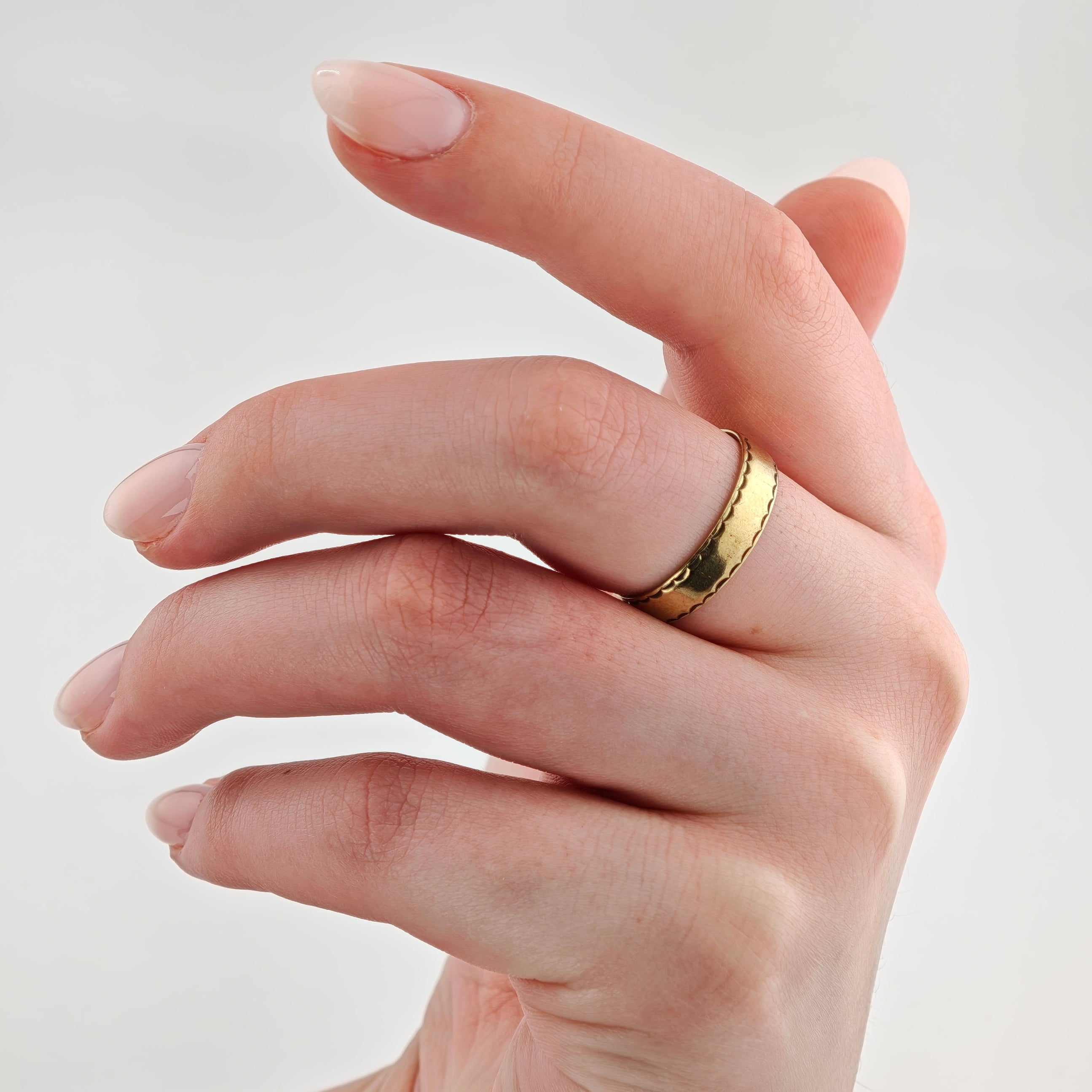 Linke Hand mit rosa lackierten Nägeln und goldfarbenem Ring mit gewellten Kanten vor neutralem Hintergrund.