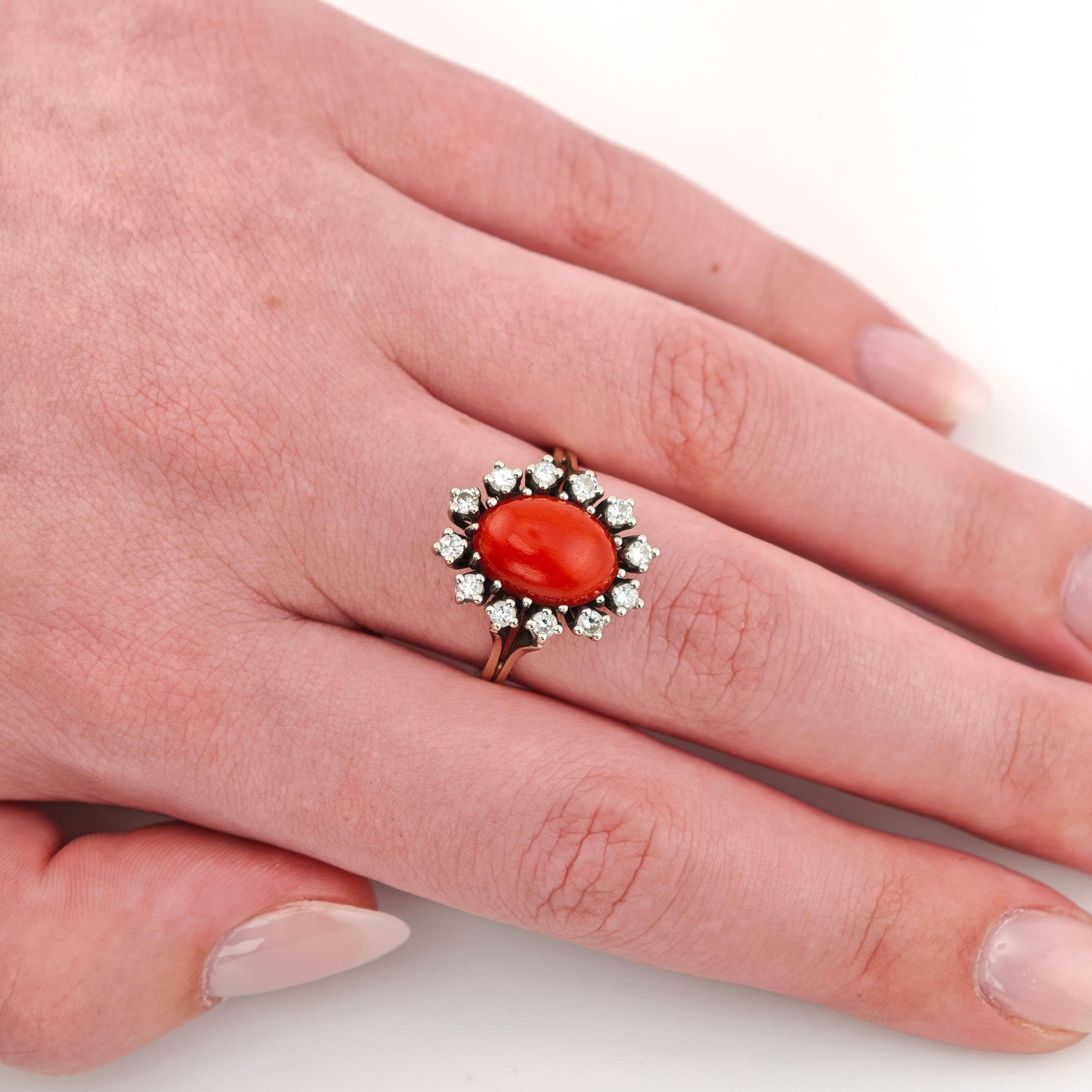 Linke Hand mit Goldring und ovalem rotem Edelstein, umgeben von kleinen klaren Steinen, vor weißem Hintergrund.