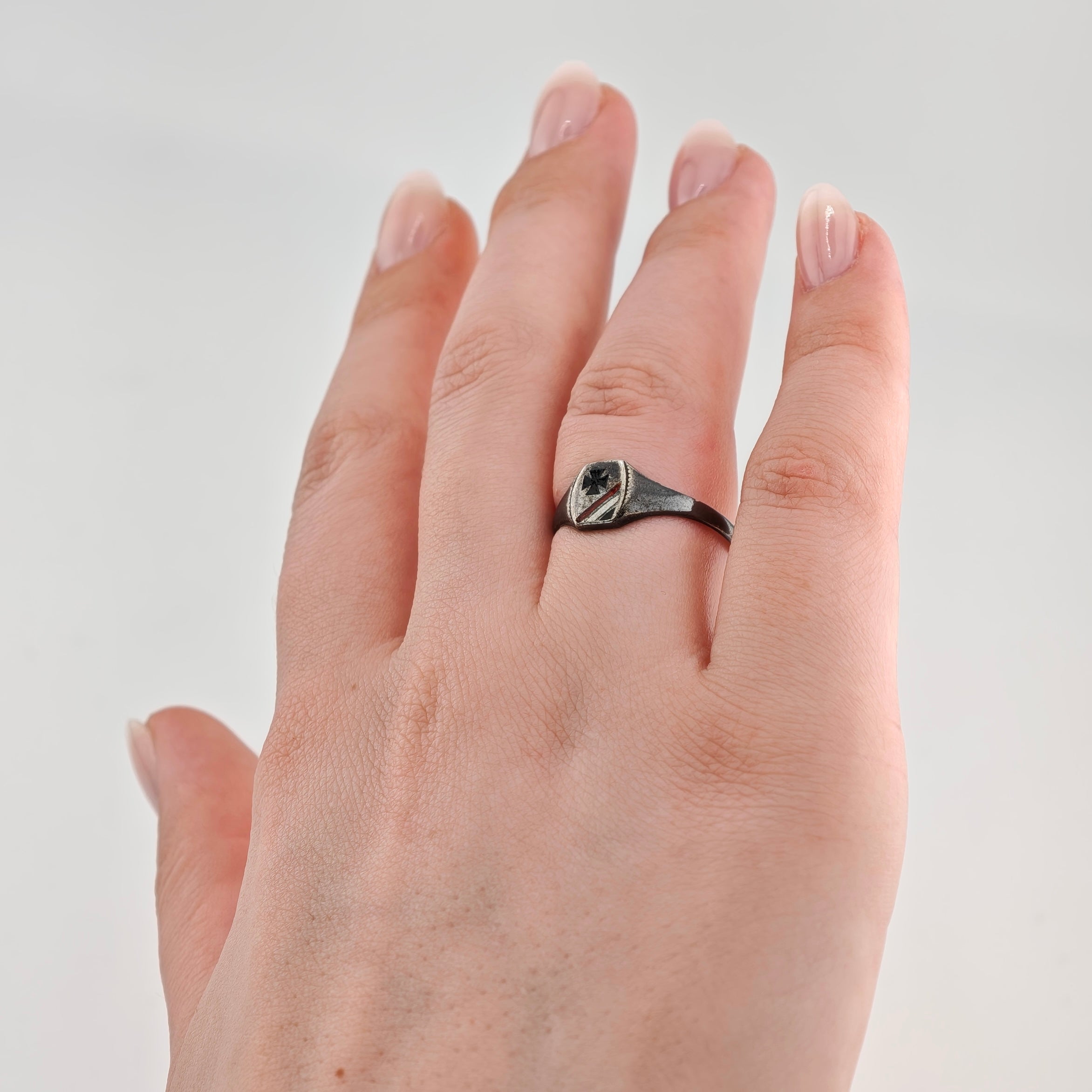 Linke Hand mit gepflegten Nägeln und schwarzem Metallschildring mit Kreuz und Streifen.