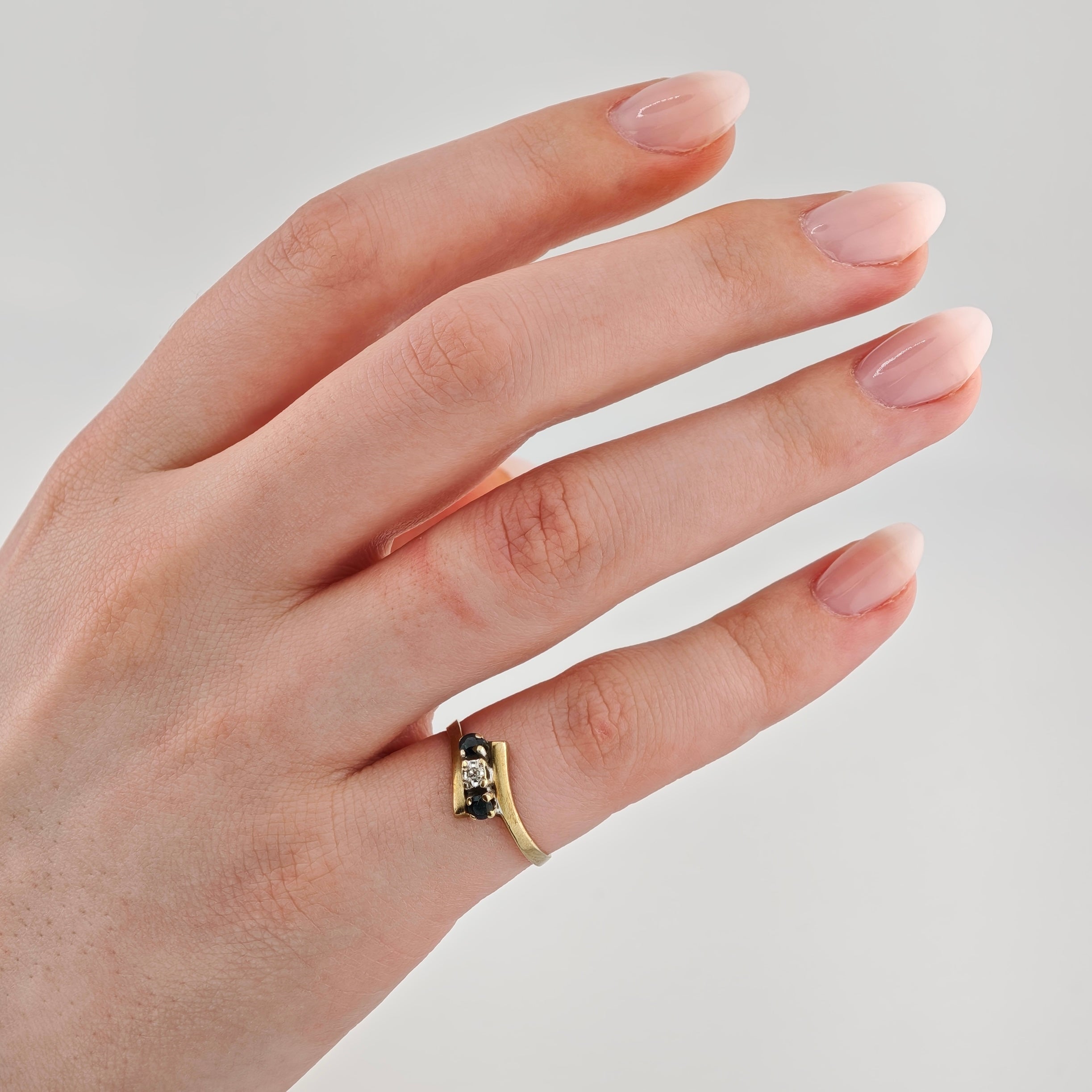 Linkerhand mit Goldring am Ringfinger mit klarem zentralen und zwei kleinen dunkelblauen Steinen vor grauem Hintergrund.