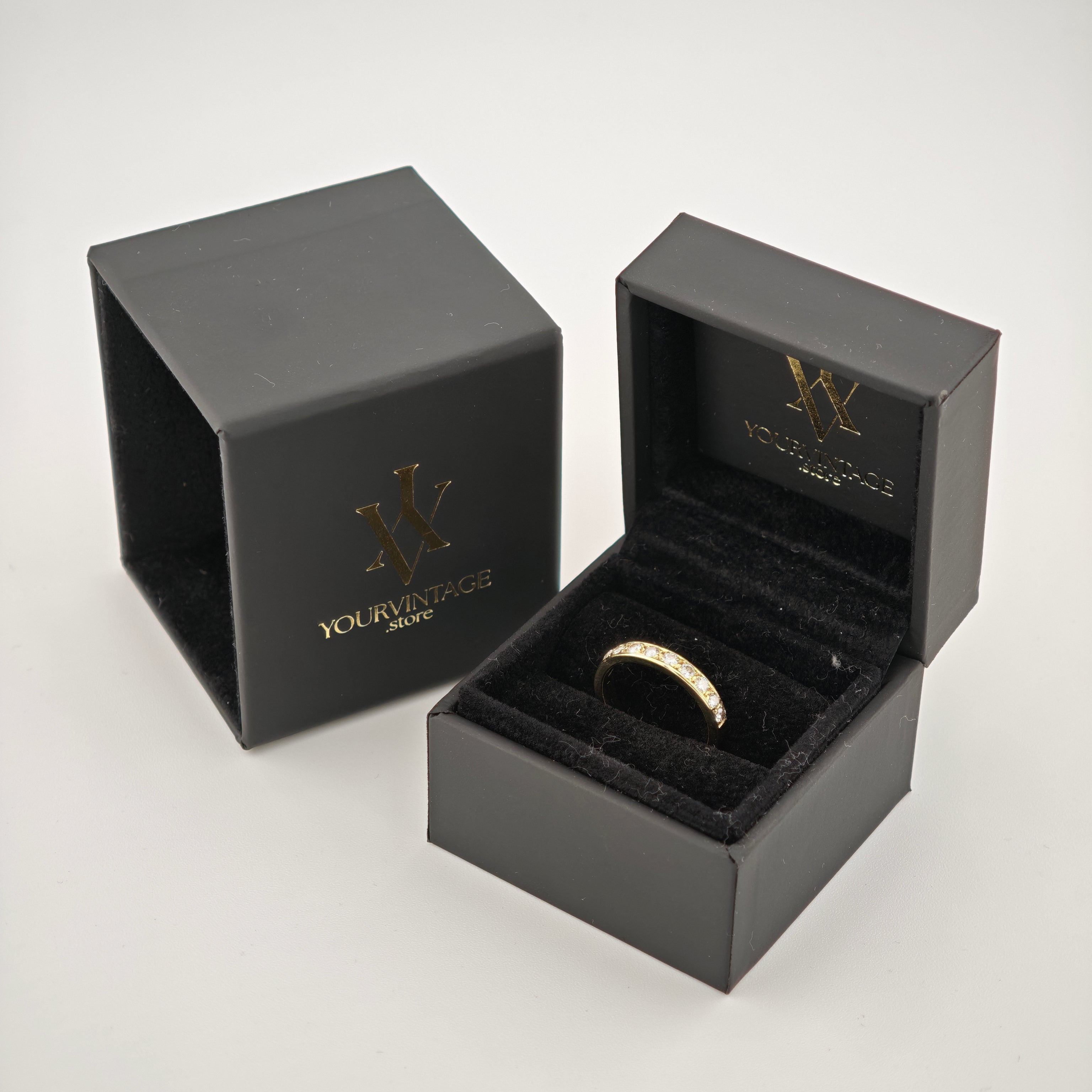 Schwarze samtige Schmuckbox mit Goldring und klaren Steinen, neben passender Verpackung mit Goldlogo auf weißem Hintergrund.