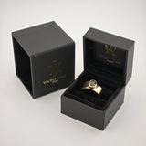 Zwei schwarze Schmuckschatullen mit goldenem Logo, eine offen mit breitem Goldring und dunklem Edelstein.