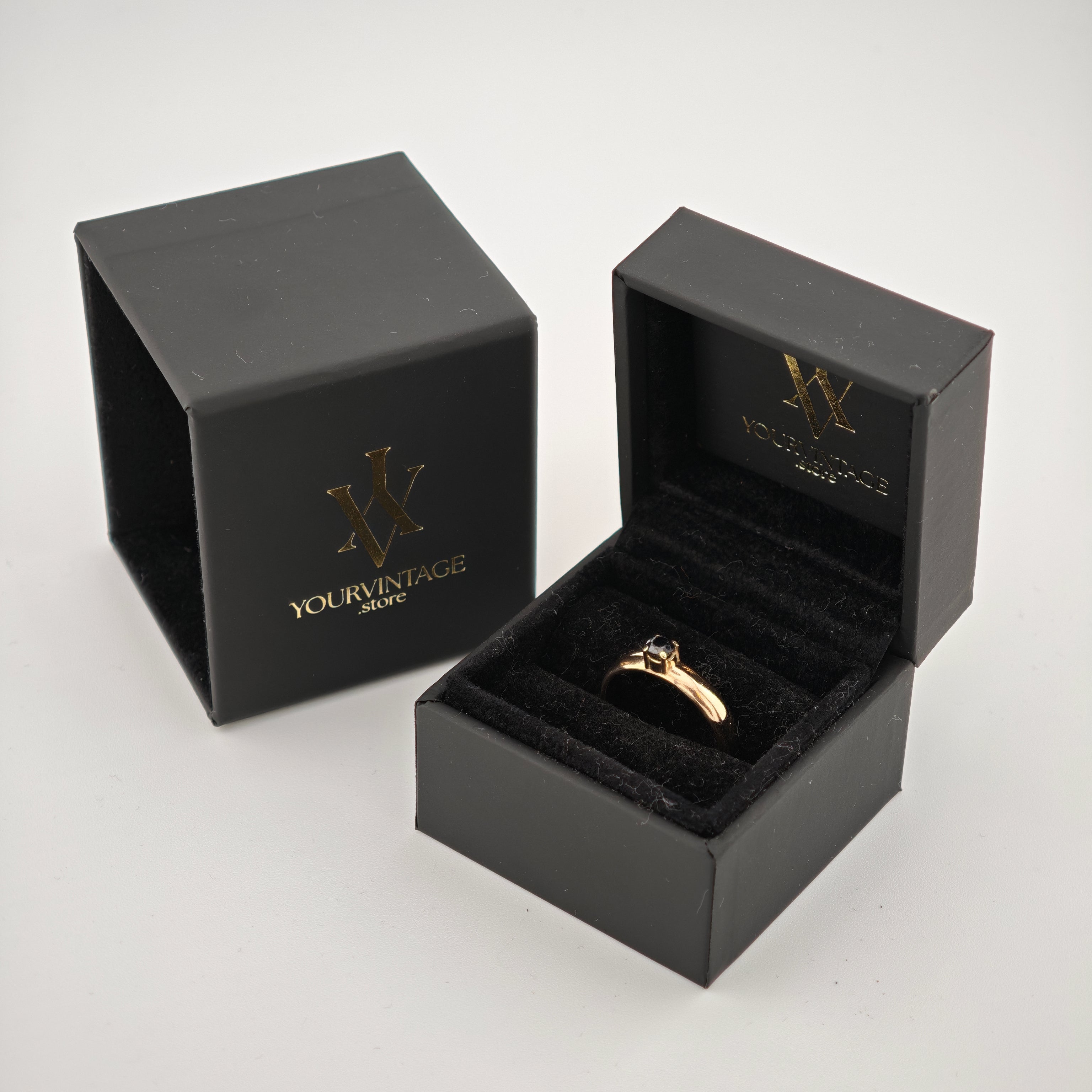 Schwarze Samtschmuckschachteln mit Goldlogo und Goldring mit dunkelblauem Stein auf weißem Hintergrund.