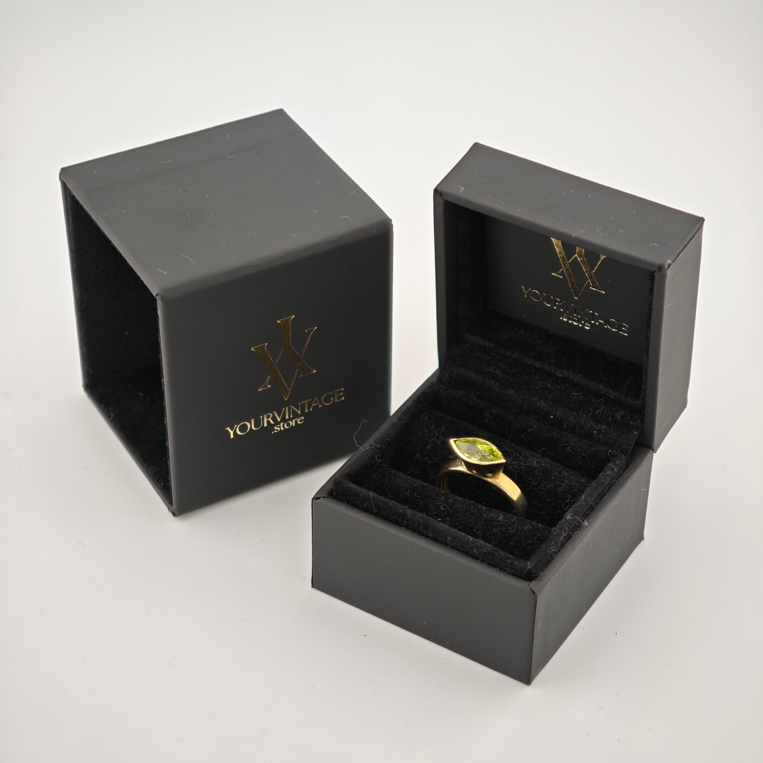 Offene schwarze Schmuckbox mit Goldring und grünem Marquise-Edelstein auf weißer Fläche.