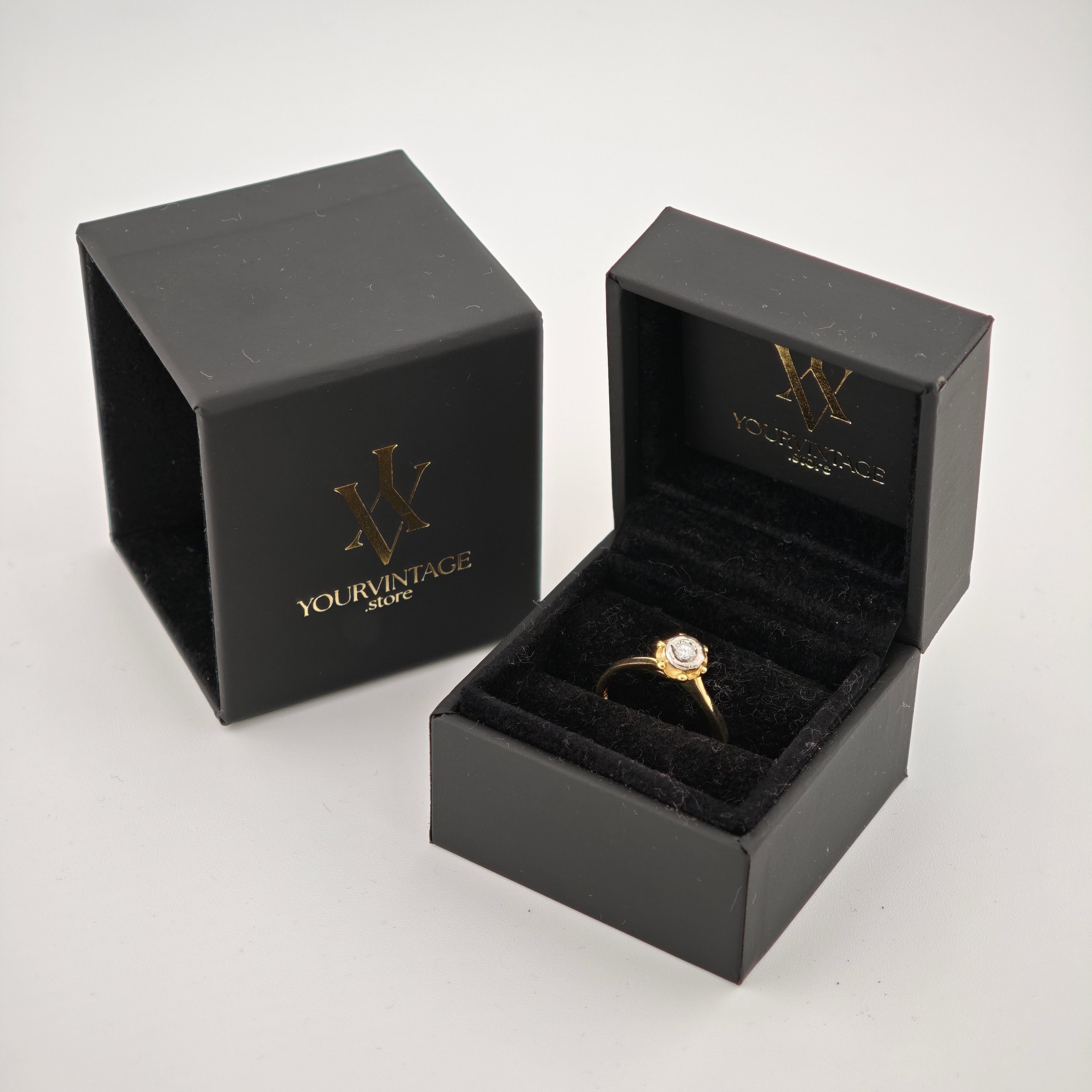 Schwarze Schmuckkästchen mit goldener Signatur und offenem Kästchen, das einen goldenen Ring mit klarem Edelstein zeigt.