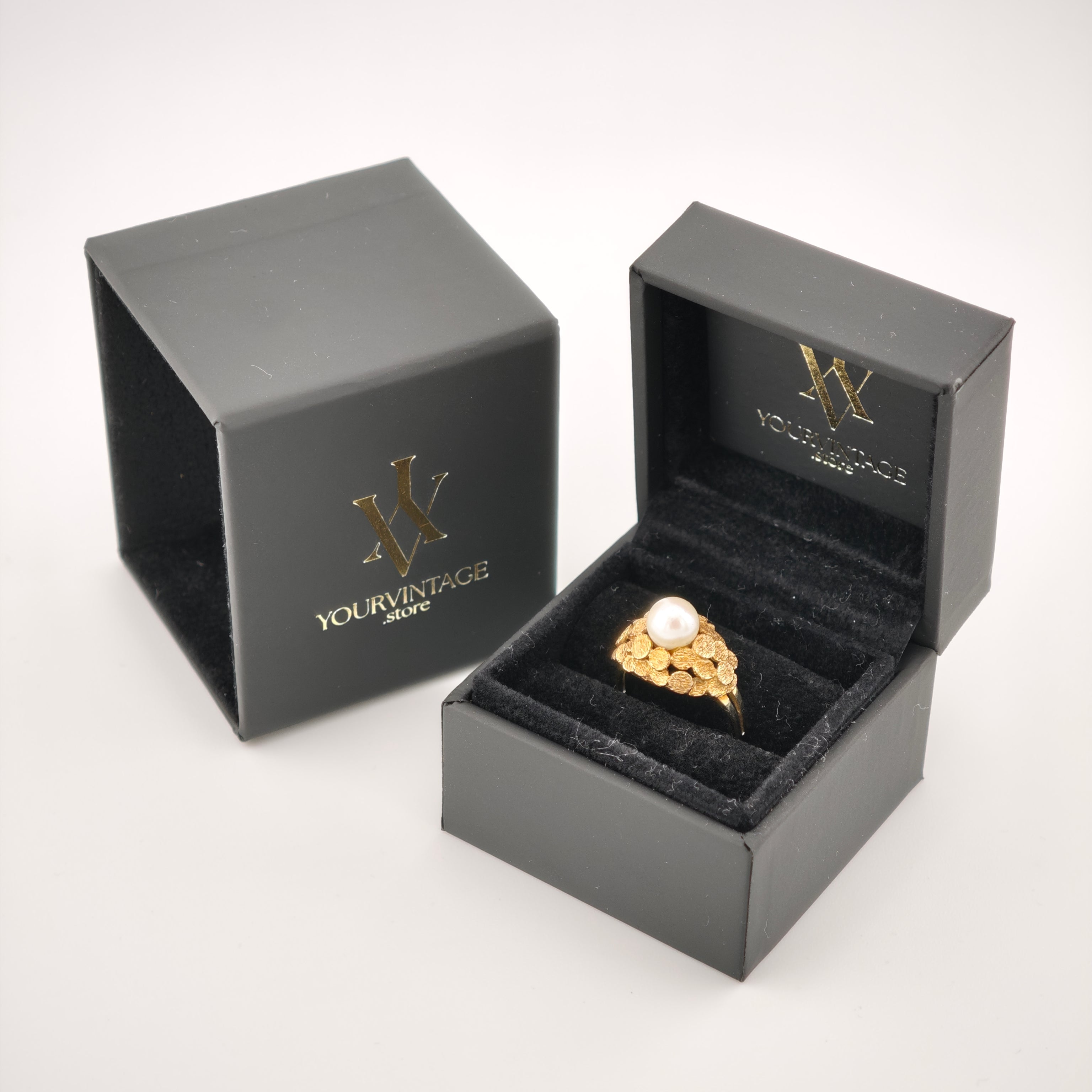 Geöffnete schwarze Schmuckschachtel mit Goldring und Perlenbesatz auf schwarzem Samt, daneben geschlossene Box mit Logo.