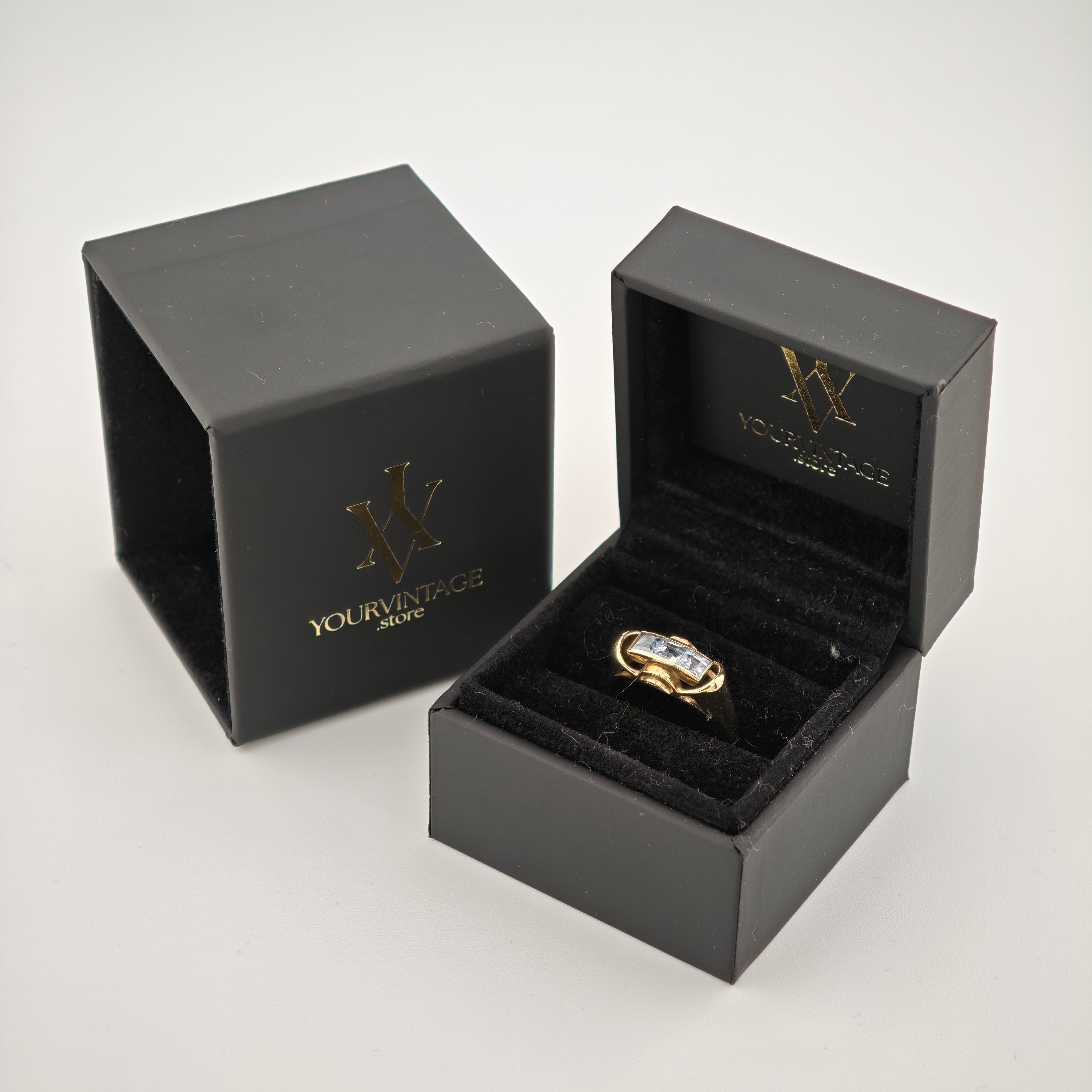 Schwarze Schmuckschachteln mit goldenem 'W'-Logo und Text 'YourVintage Store', offene Schachtel zeigt goldenen Ring mit drei klaren rechteckigen Edelsteinen.