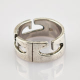 Silberner offener Ring mit stilisierten G-förmigen Ausschnitten und texturierter Oberfläche auf reflektierendem Hintergrund.