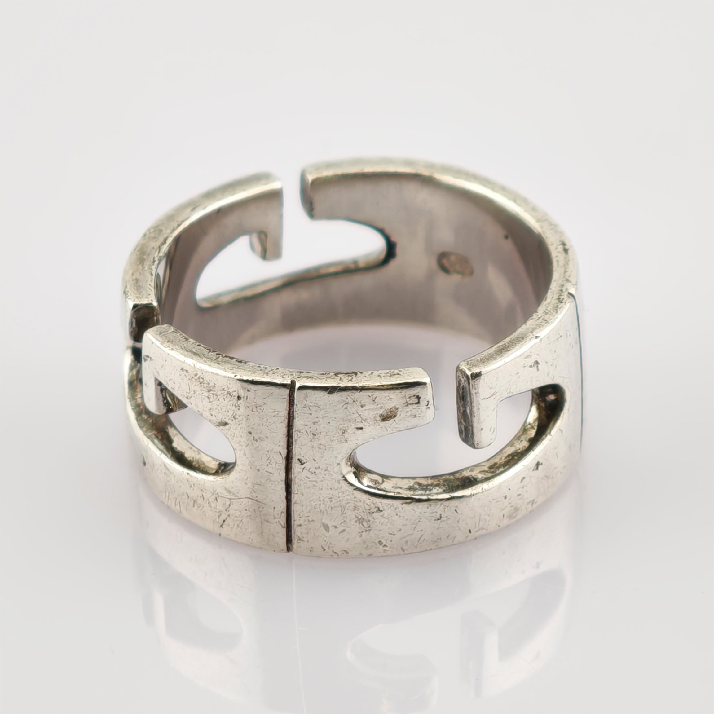 Silberner offener Ring mit stilisierten G-förmigen Ausschnitten und texturierter Oberfläche auf reflektierendem Hintergrund.