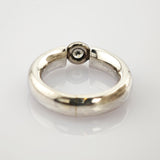 Silberner Ring mit glatter Oberfläche und schwarzem rundem Detail auf weißem reflektierendem Hintergrund.