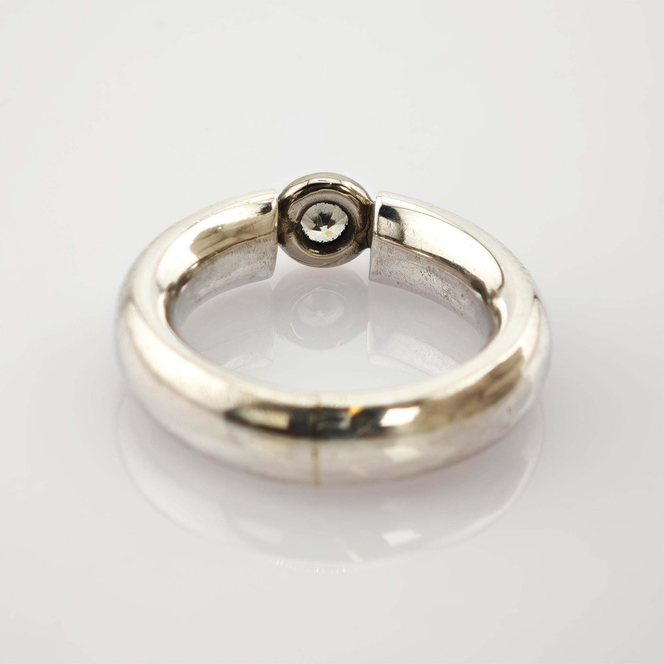 Silberner Ring mit glatter Oberfläche und schwarzem rundem Detail auf weißem reflektierendem Hintergrund.