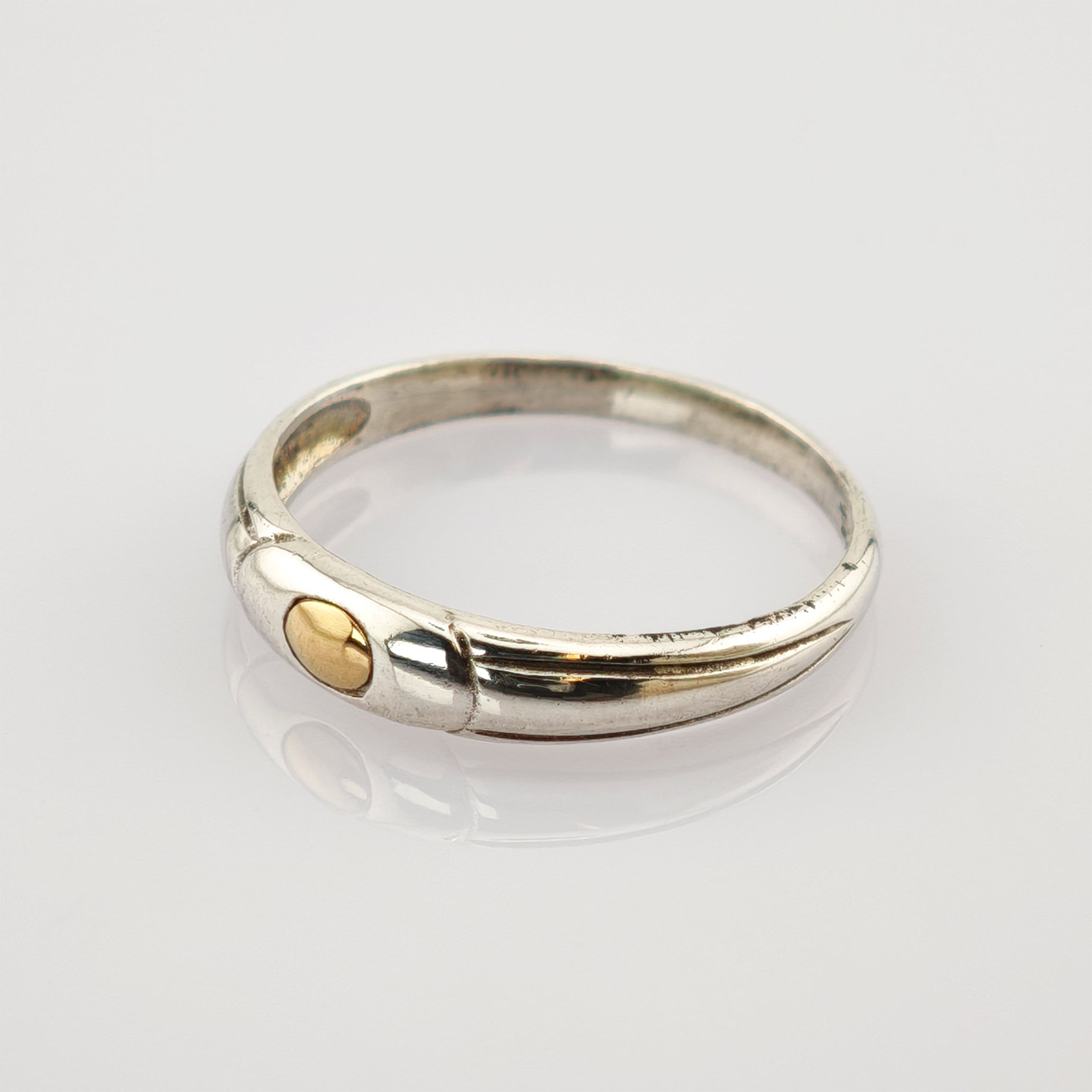 Silberner Ring mit kleinem goldfarbenem, kreisförmigem Detail auf reflektierender, weißer Oberfläche.