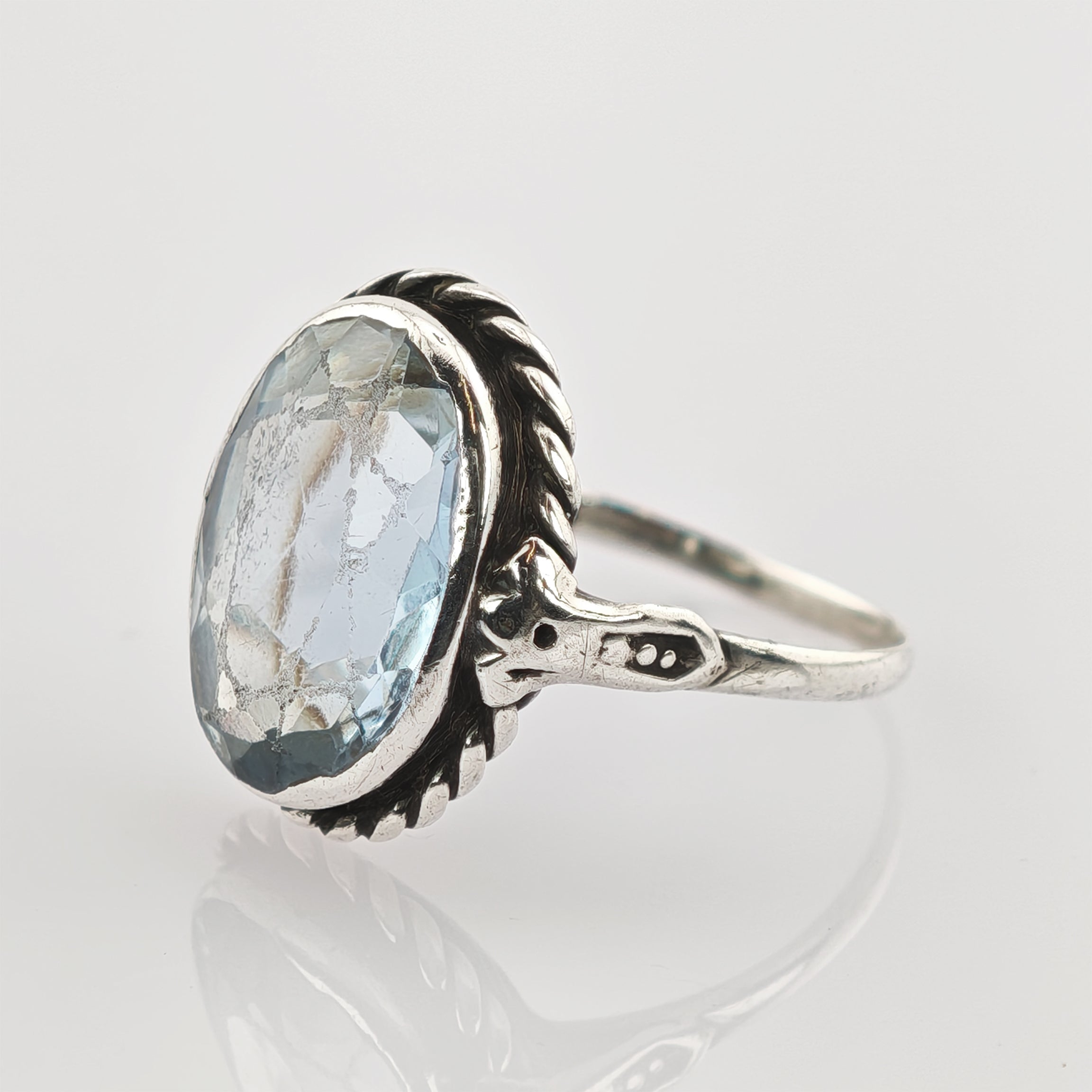 Nahaufnahme eines silbernen Rings mit ovalem, hellblauem, facettiertem Edelstein und kunstvollem Seilrand.