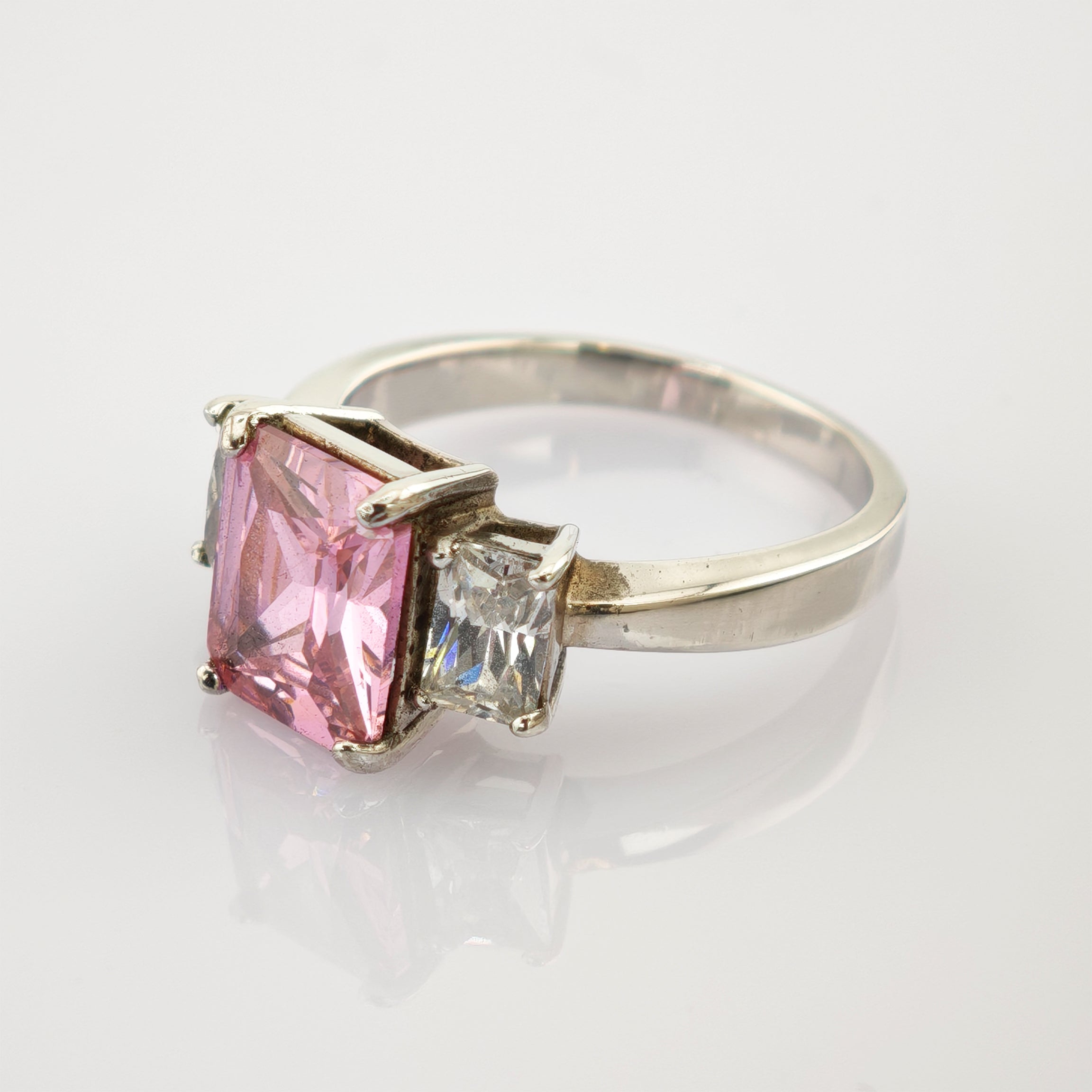 Silberner Ring mit rechteckigem rosa Edelstein und klarem Edelstein auf reflektierender Oberfläche.