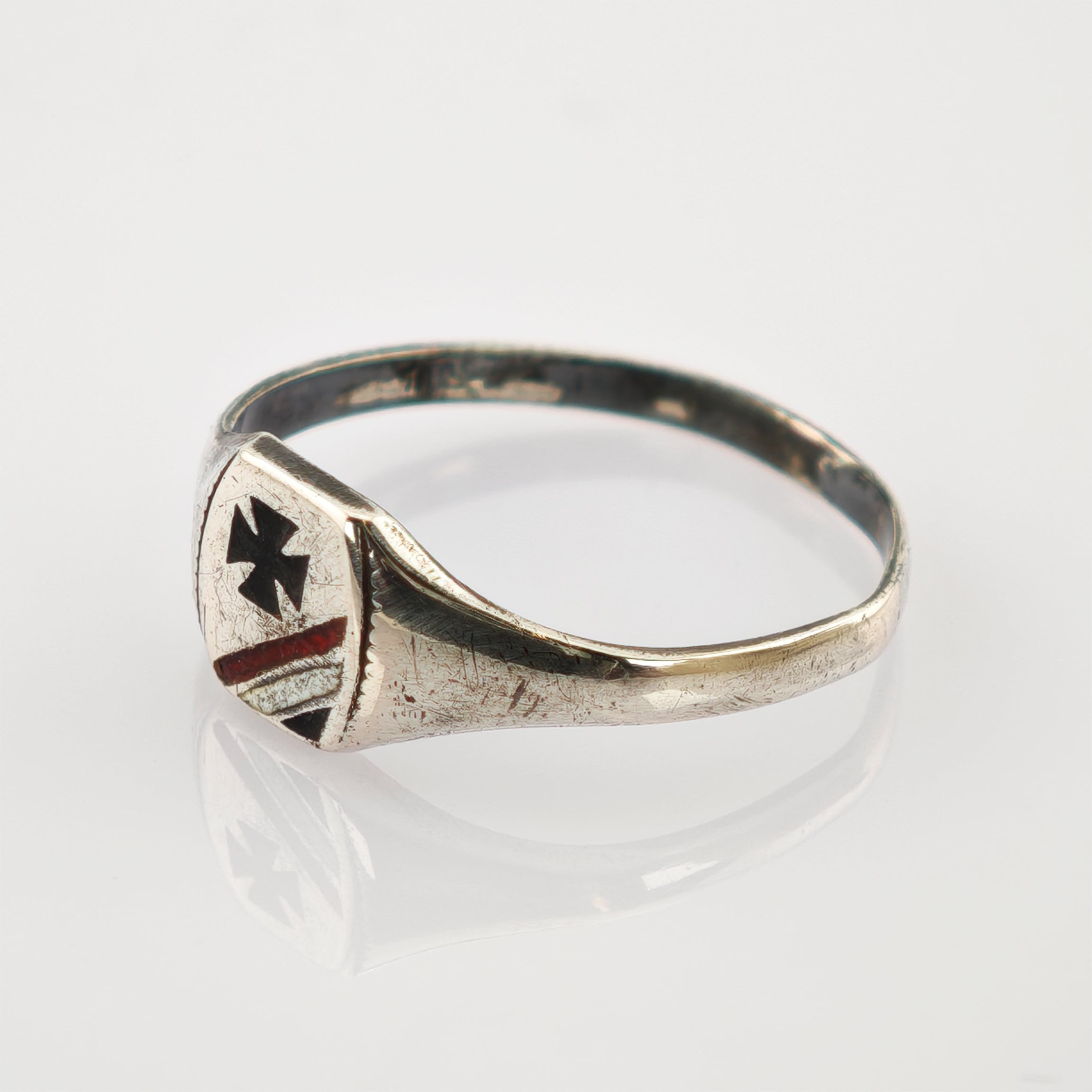 Tarnierter silberner Ring mit schildförmiger Platte, schwarzem Kreuz und rot-silbernen Streifen auf weißer Oberfläche.