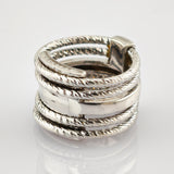 Nahaufnahme eines silbernen Rings mit verflochtenen, gedrehten Bändern und glattem Mittelband auf reflektierender Oberfläche.