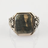 Silberfarbener Signet-Ring mit rechteckigem, facettiertem Gesicht und floralen Verzierungen auf weißem Hintergrund.